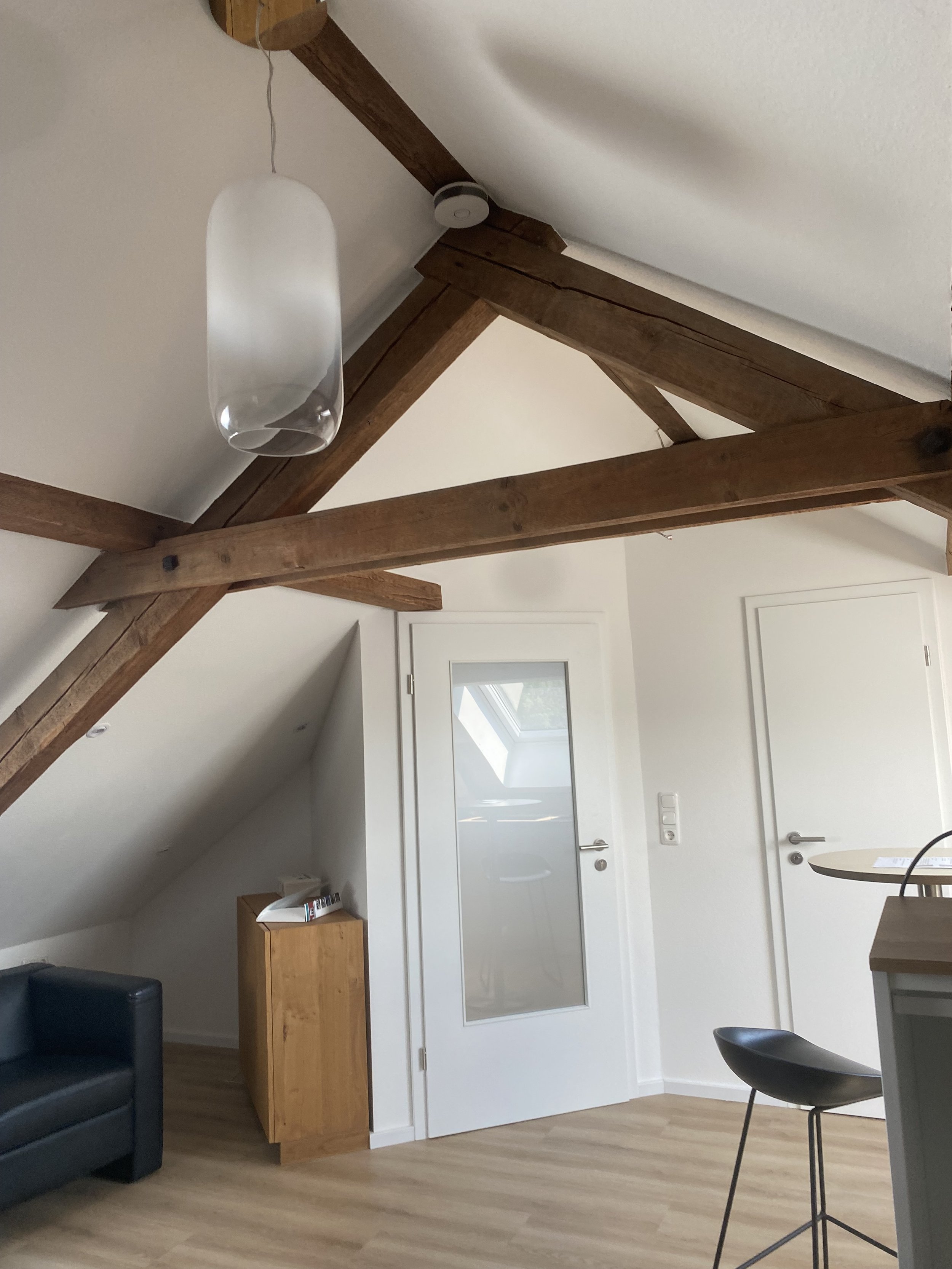 Innenraum eines Zimmers mit Holzbalken an der Decke, einer weißen Tür mit Glasfenster, einem schwarzen Sofa, einem kleinen Holzschrank mit Telefon, einer modernen Deckenlampe und einem schwarzen Stuhl.