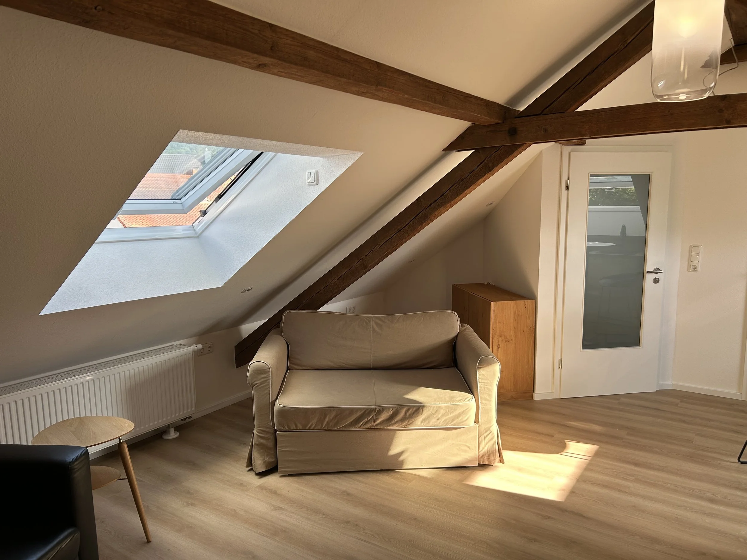 Ein heller Raum mit Holzboden, couch, Sprossenfenster in der Dachschräge, Holzbalken an der Decke, weiße Wände, Glastür, kleine Holzkommode.