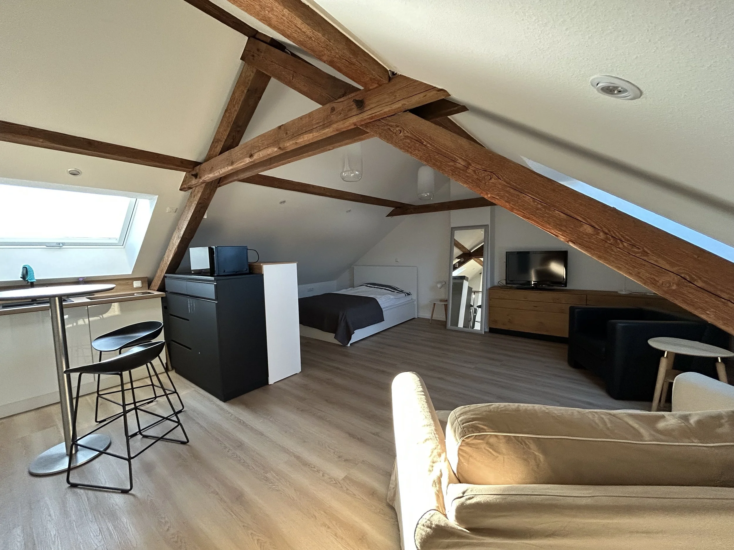 Gemütliches Schlafzimmer im Dachgeschoss mit Holzträgern, ein Doppelbett, ein Fernseher auf einem Lowboard, ein spiegel und eine Couch, viel natürlicher Lichteinfall durch Dachfenster.