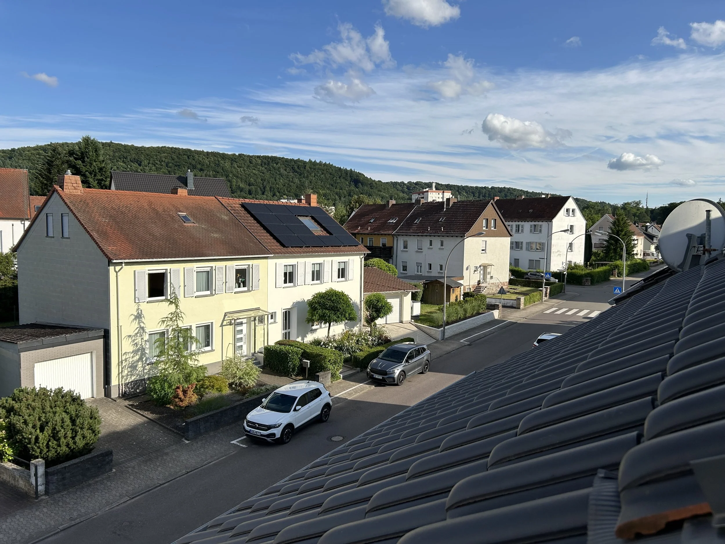 Blick auf ein Wohnviertel bei sonnigem Himmel mit einigen Wolken, mehrere Häuser mit Gärten und parkenden Autos, hinter den Gebäuden eine grüne Hügelkette.