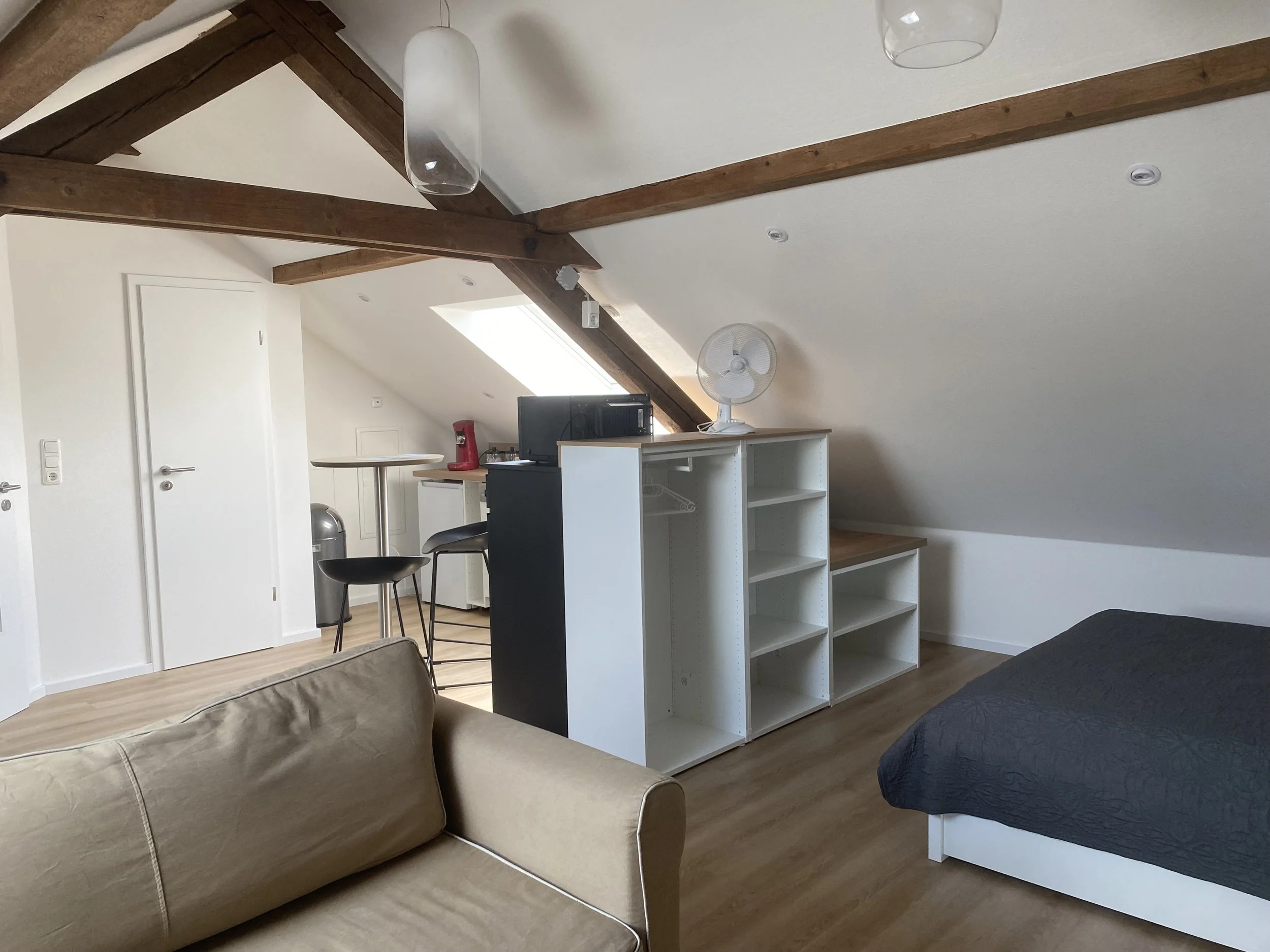 Ein Schlafzimmer mit schrägen Decken und sichtbaren Holzbalken, inklusive einem Bett, einem Sofa, einem Schreibtisch, einem Fernseher, einem Ventilator, einer kleinen Küche mit Kaffeemaschine, einer Spüle, einem Barhocker und einem Staubsauger.