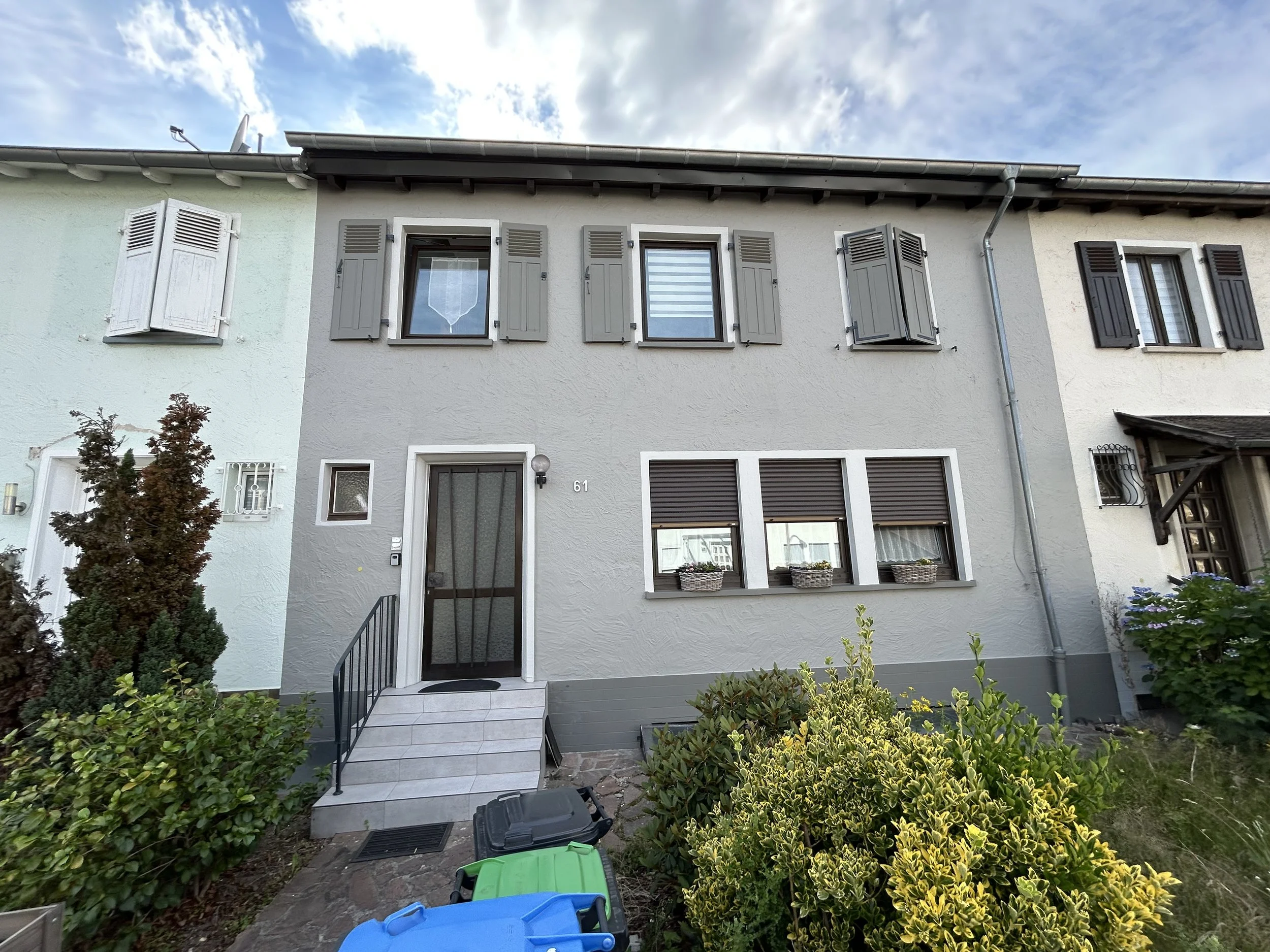 Frontansicht eines zweistöckigen Hauses mit weißen und grauen Wänden, mehreren Fenstern mit Fensterläden, Haustür mit Gitter und kleinen Stufen, umgeben von Büschen und Pflanzen, unter einem bedeckten Himmel.