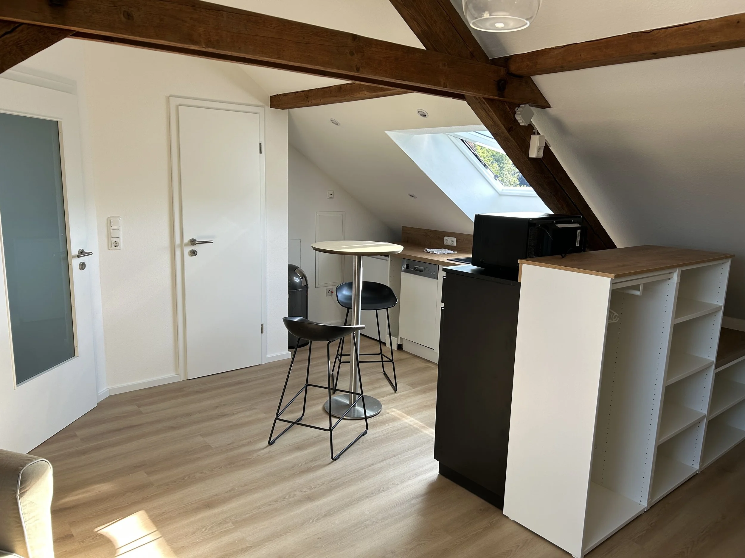 Ein kleines Zimmer mit Holzfußboden, weißen Wänden und Dachbalken. Es gibt eine kleine Küchenzeile mit Mikrowelle, Geschirrspüler und zwei schwarzen Barhockern. Ein Fenster an der Dachschräge lässt Tageslicht herein.