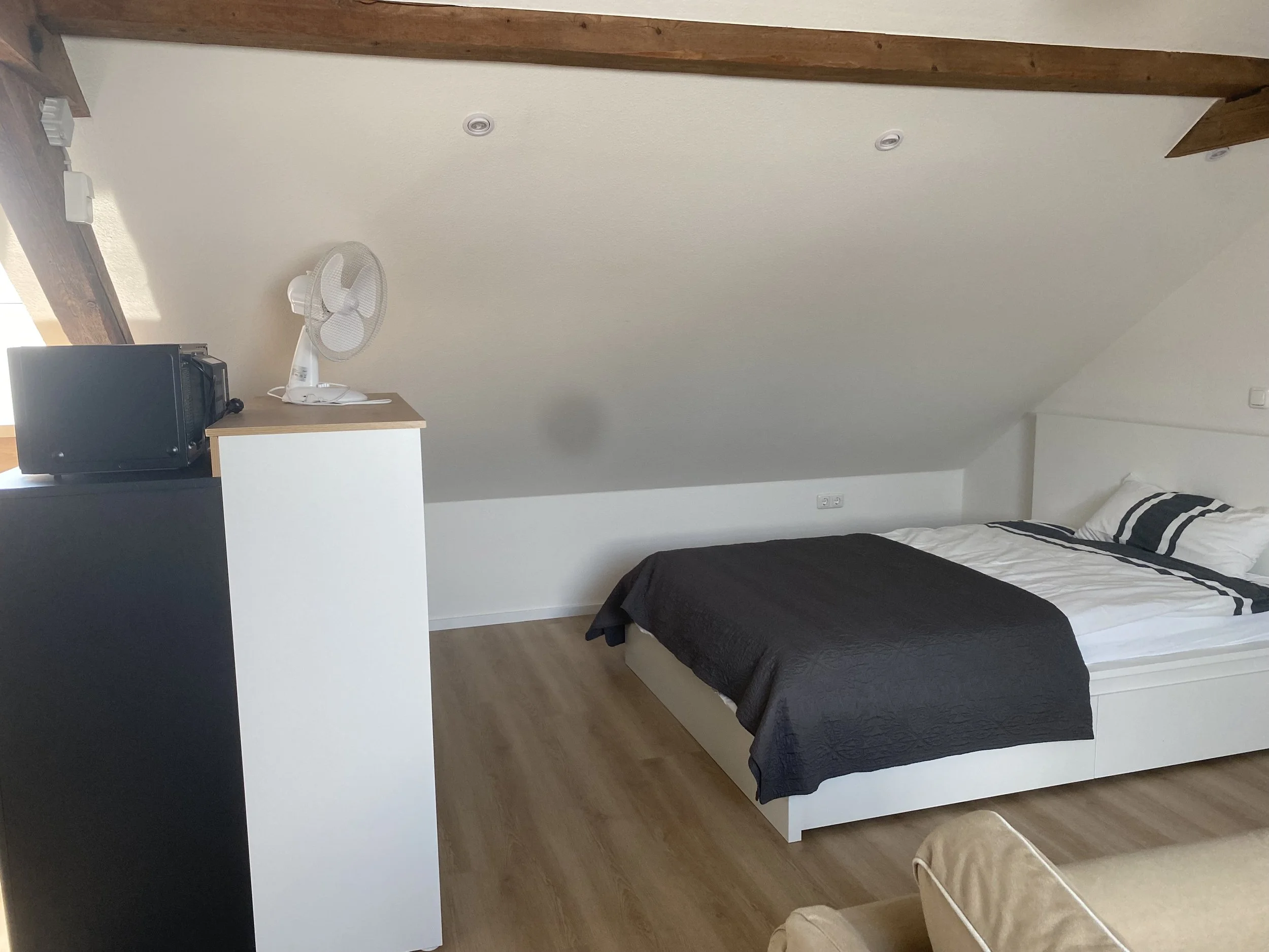 Schlafzimmer mit weißem Bett mit schwarzen Decke und Kissen, kleinen Fenstern, Holzboden, einem kleinen weißen Schrank mit Mikrowelle und Ventilator, weiße Wände und Dachschräge.