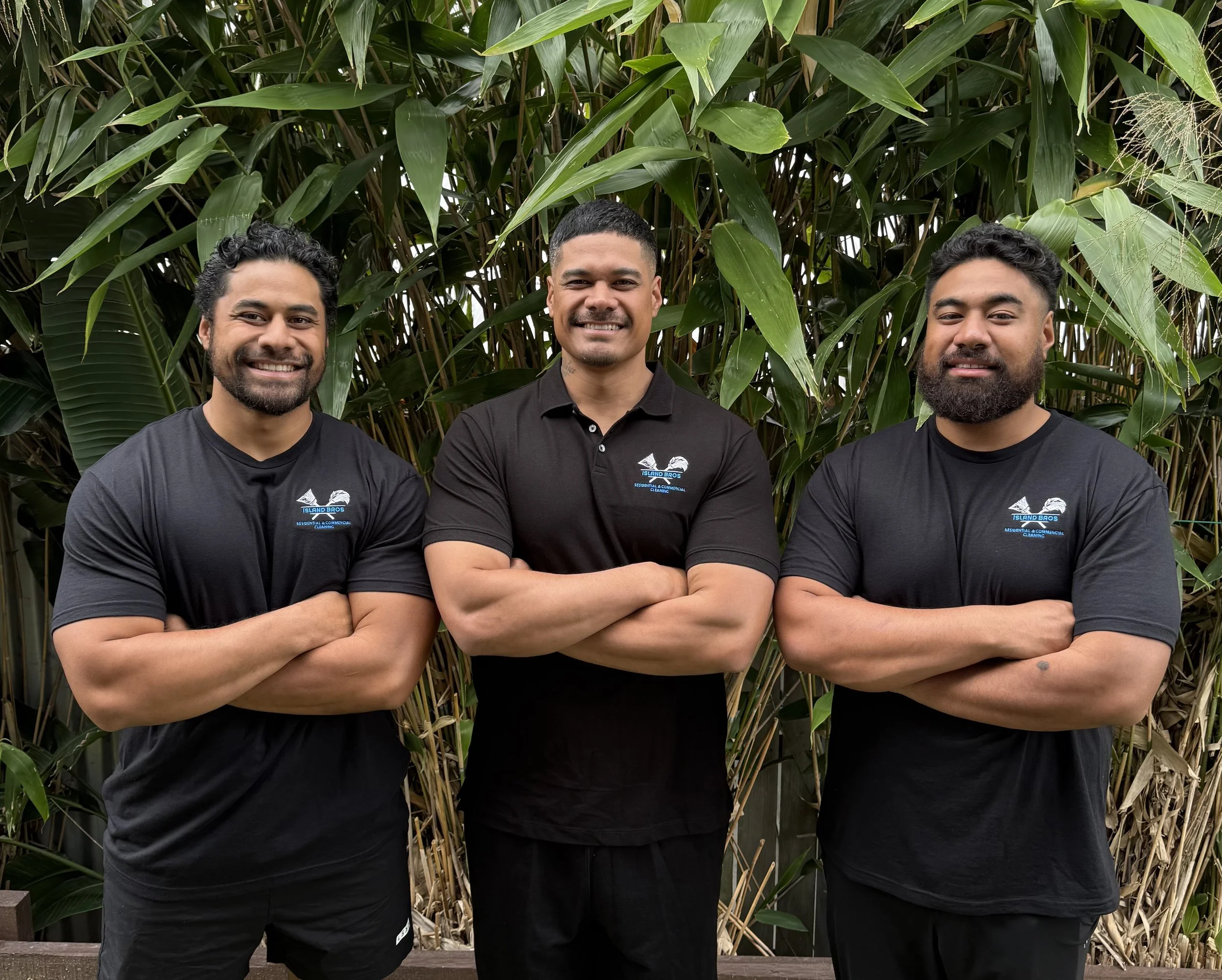 Three men standing outdoors in front of lush green plants with crossed arms, smiling at the camera, wearing black t-shirts with a logo.