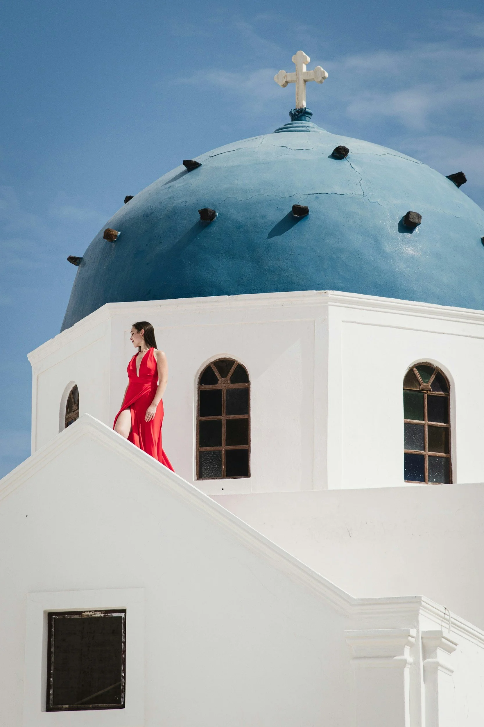 Iconic Blue dome Santorini. A woman in a red dress standing on a white building with a blue domed roof and a cross on top, against a blue sky.