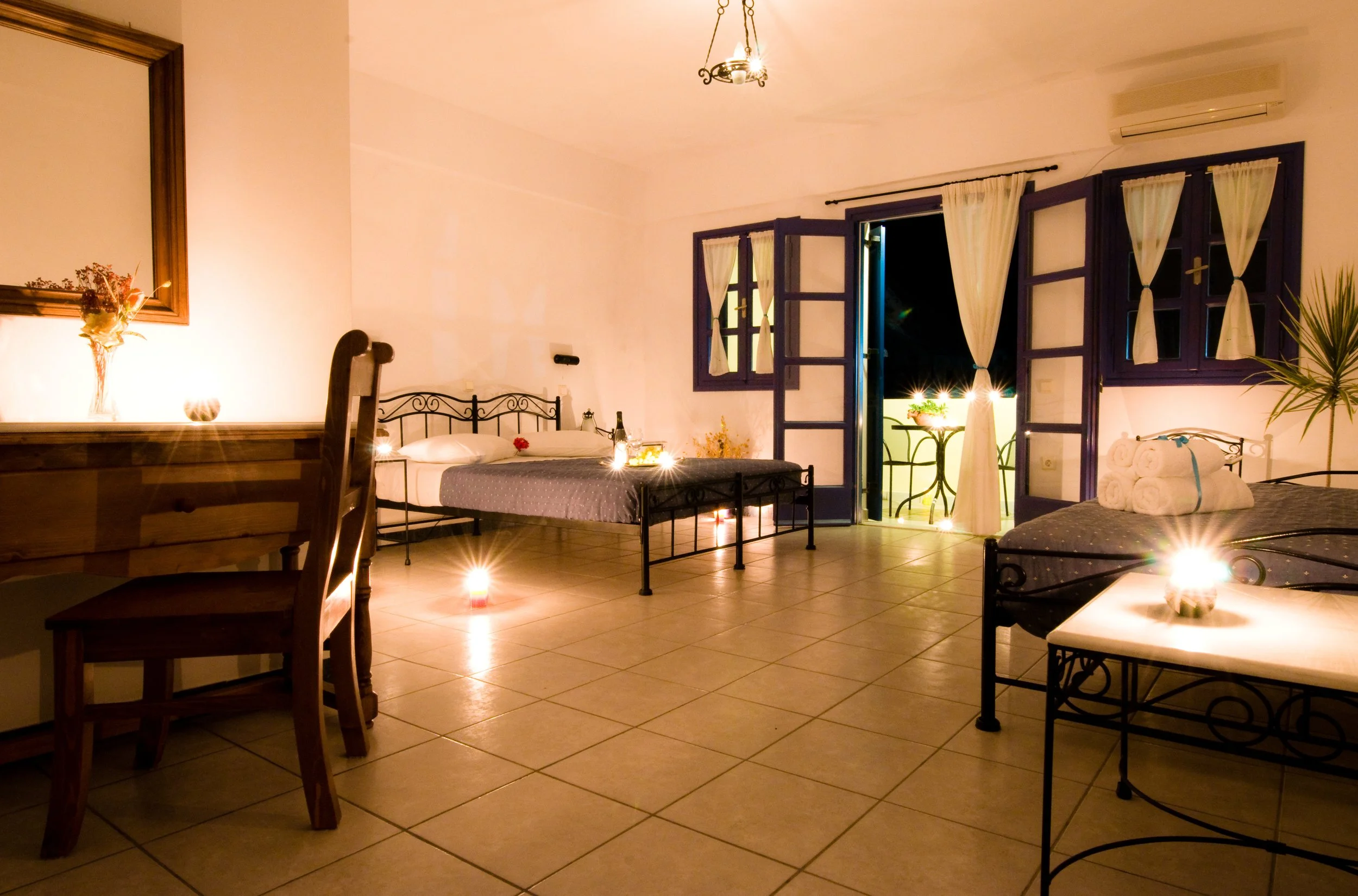 A warmly lit bedroom with two beds, an open balcony door, candles, towels, and a dining table outside.