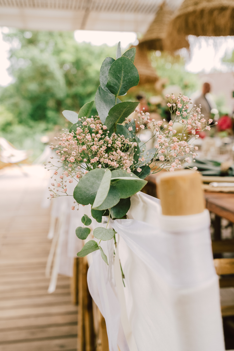 Blumenarrangement mit grünen Blättern und kleinen rosa Blüten an einer dekorierten Holzbank im Freien.