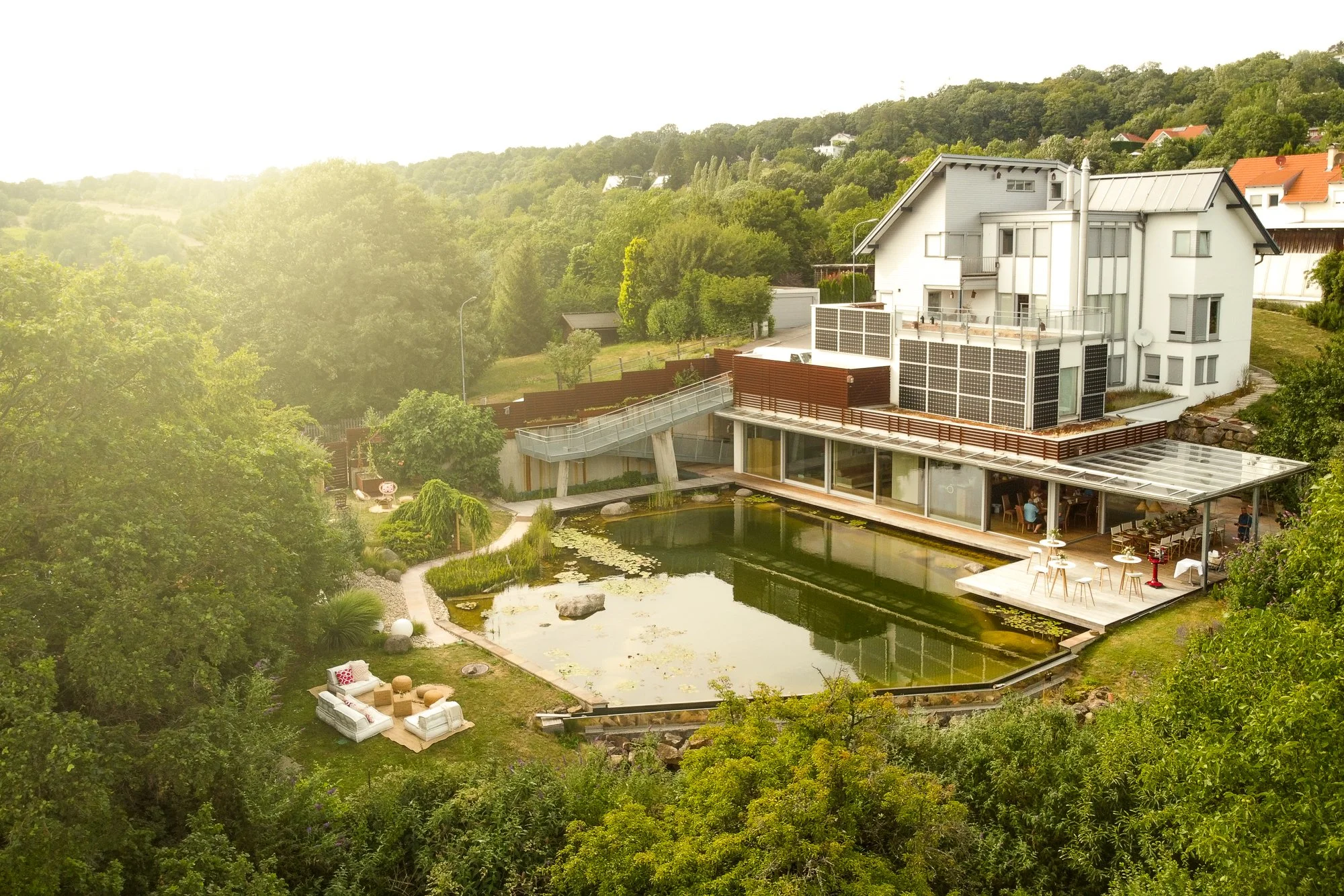 Moderne weiße Villa mit Terrasse und Teich, umgeben von grünen Bäumen, in ländlicher Umgebung.