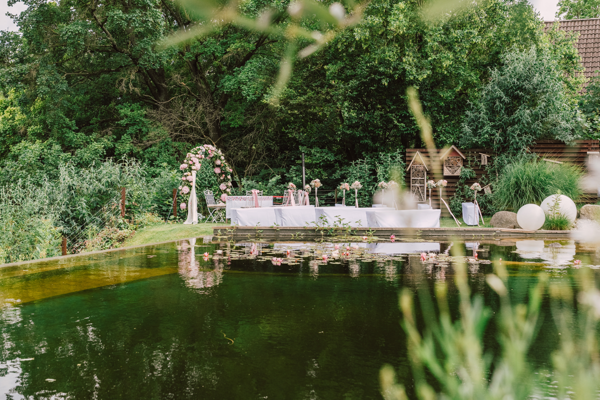 Ein schön gedeckter Tisch im Garten, umgeben von Bäumen und Pflanzen, mit einem Wasserbecken im Vordergrund, vermutlich für eine Feier oder Hochzeit vorbereitet.