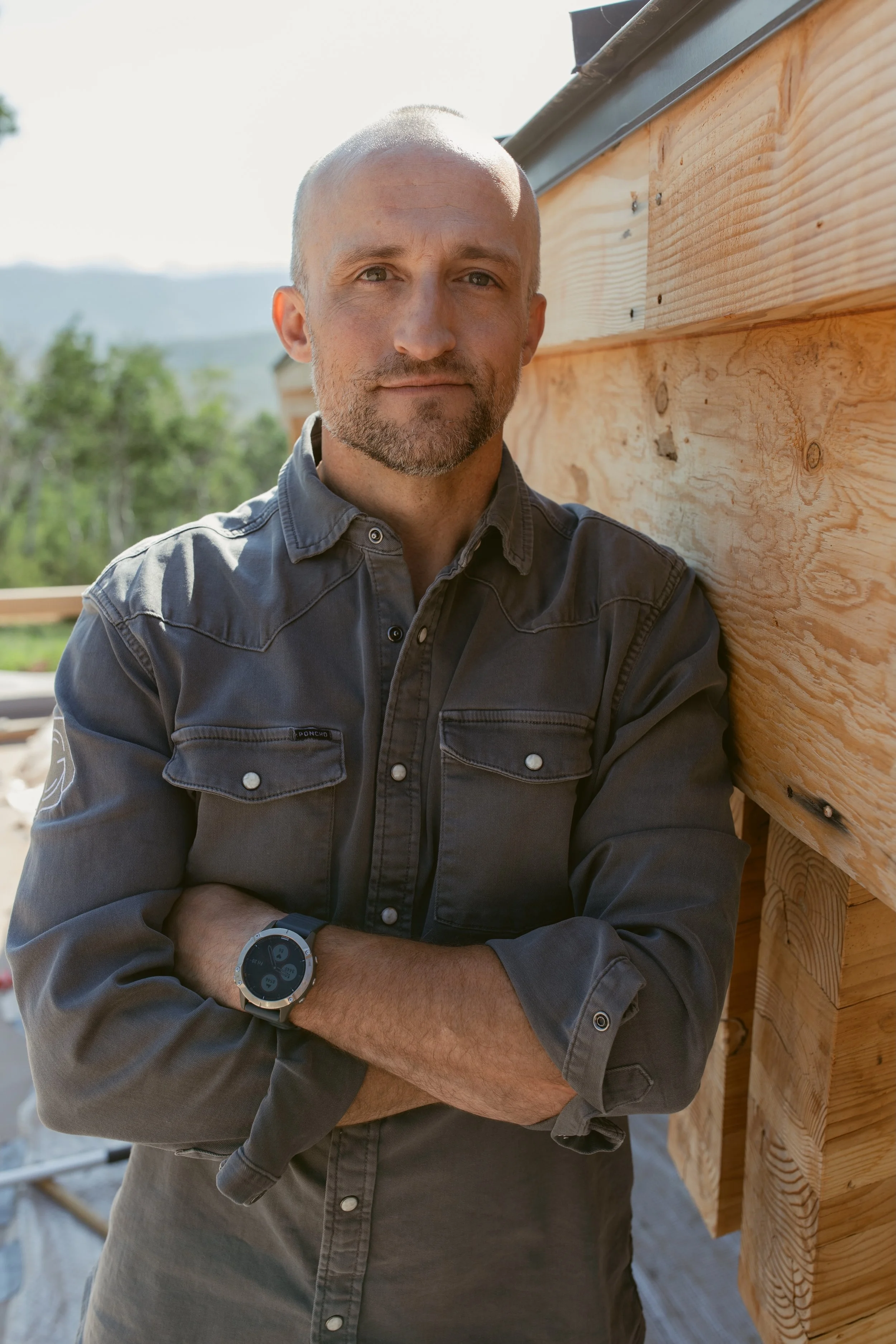 A man with a beard, wearing a gray button-up shirt and a smartwatch, stands with arms crossed next to a wooden siding on a building under construction, outdoors with green trees and mountains in the background.