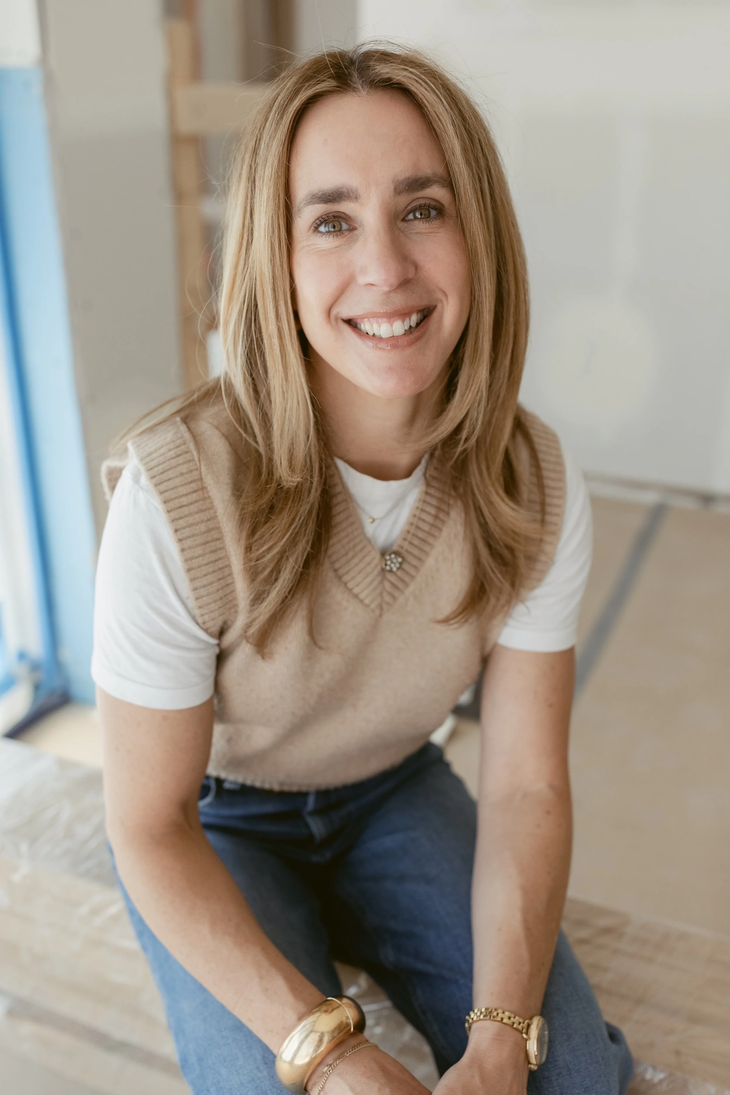 A woman with shoulder-length light brown hair, smiling, wearing a beige vest over a white t-shirt, sitting on a wooden surface in an indoor setting with a blurred background.
