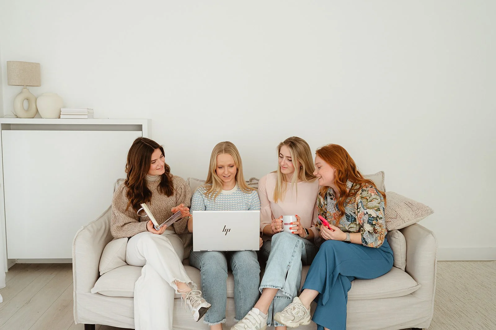 Vijf jonge vrouwen zitten op een witte bank in een minimalistische woonkamer en kijken naar een laptop.