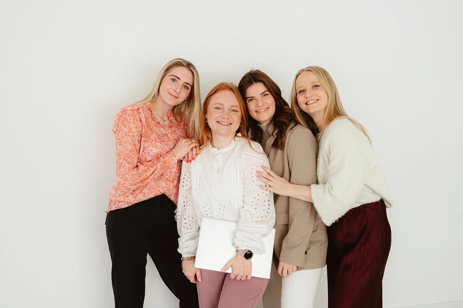 Vijf vrouwen staan samen en lachen tegen een witte achtergrond.