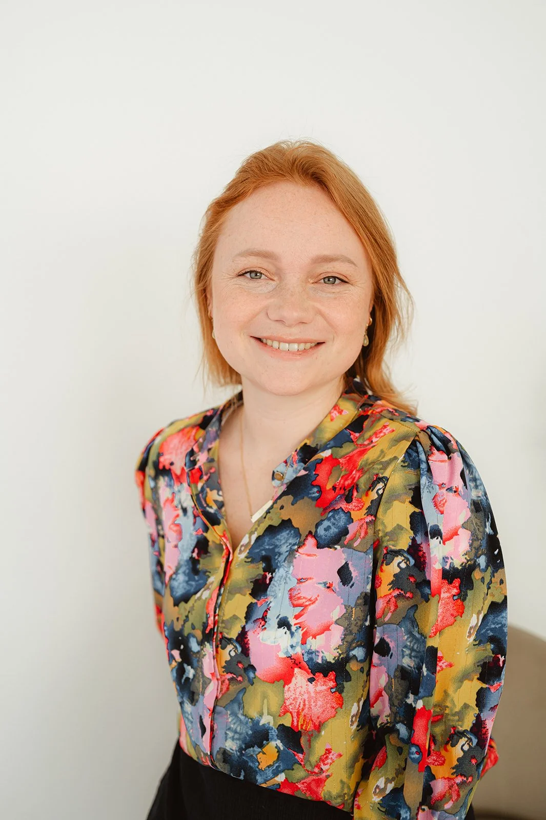 Portret van een vrouw met rood haar, bloemenpatroon blouse, lichte huid en glimlach, witte achtergrond.