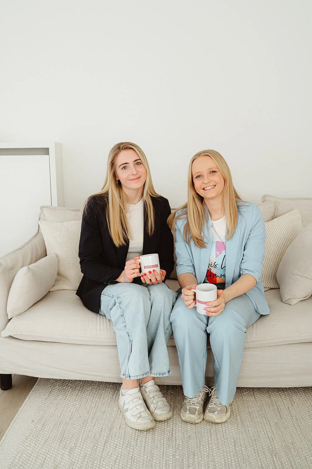 Twee vrouwen zitten samen op een bank met kopjes in een witte kamer.