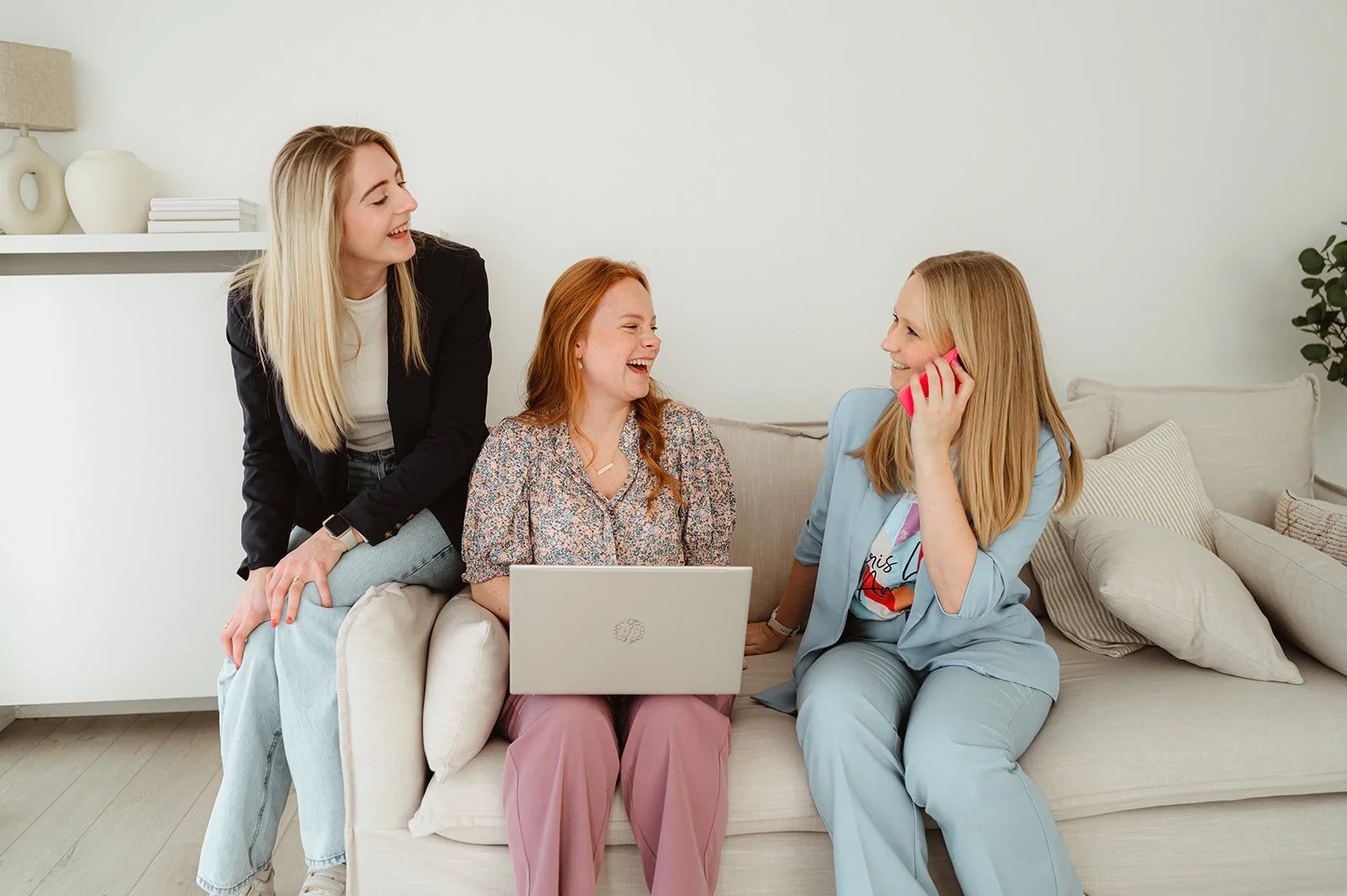 Vier vrouwen zitten op een bank in een lichte woonkamer, lachen en praten met elkaar. Eén vrouw heeft een laptop op haar schoot, een ander praat op de telefoon.