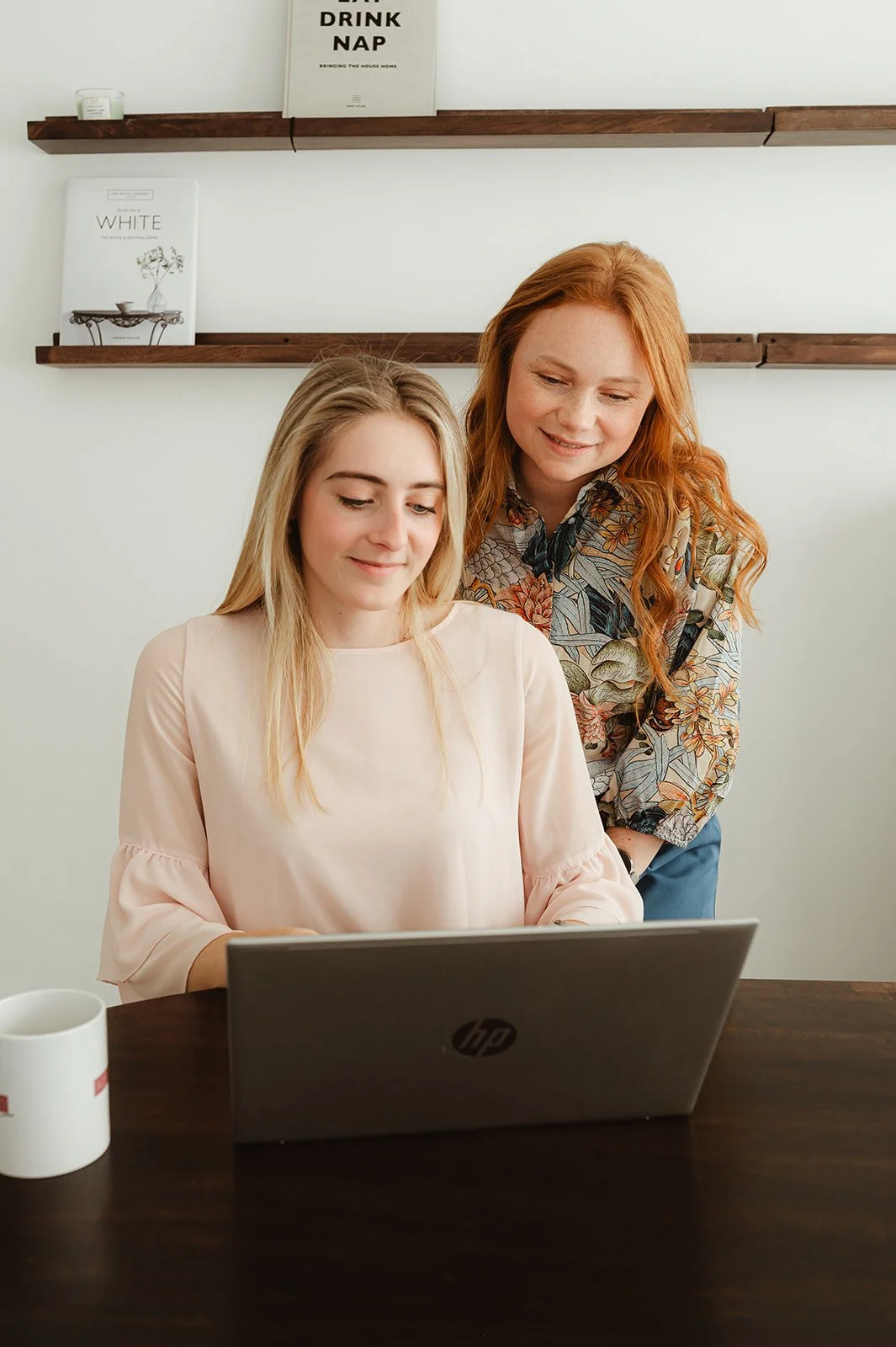Twee vrouwen kijken naar een laptop op een bureau in een moderne kamer met witte muren en houten planken.