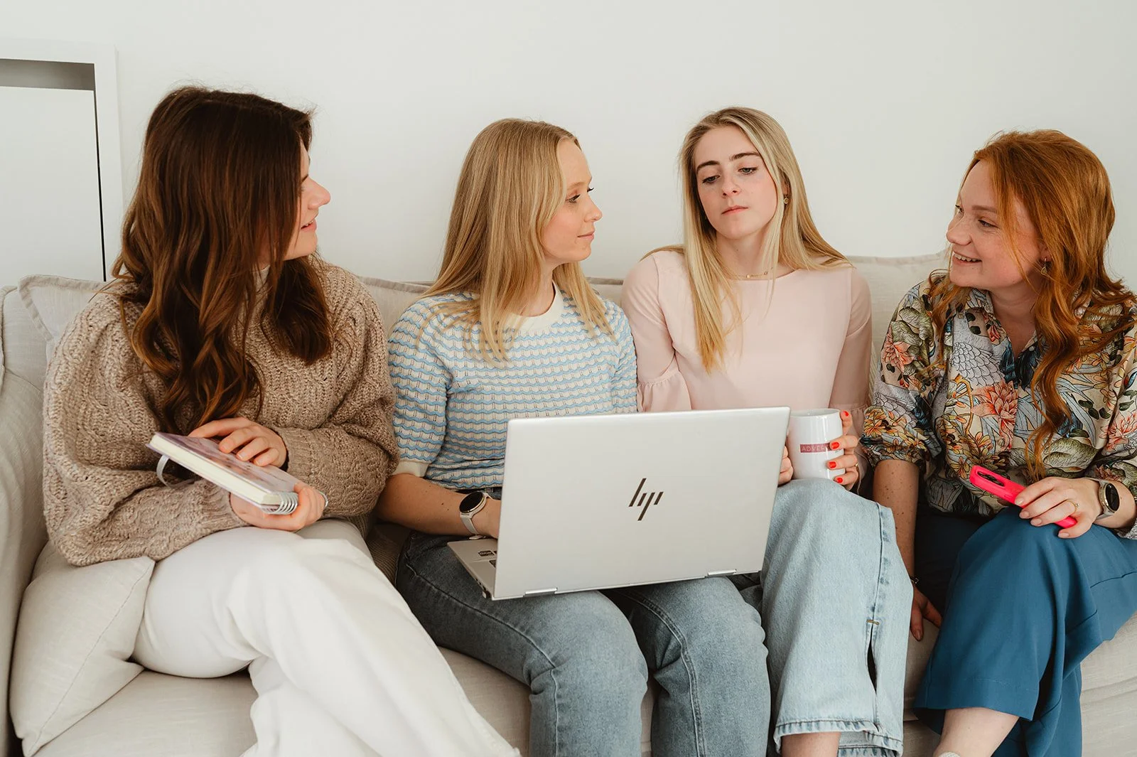 Vijf vrouwen zitten op een bank en kijken naar een laptop, terwijl ze praten en lachen.