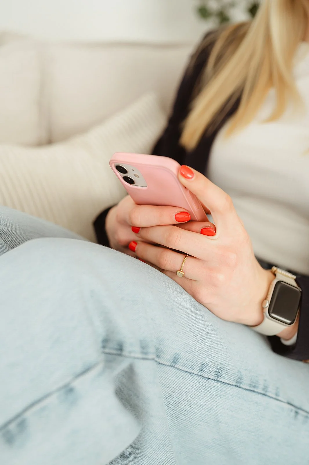 Een persoon houdt een roze smartphone vast, draagt een smartwatch en een ring met een steen. Ze zitten in een comfortabele setting met lichte kussens en kleding van lichte kleuren.