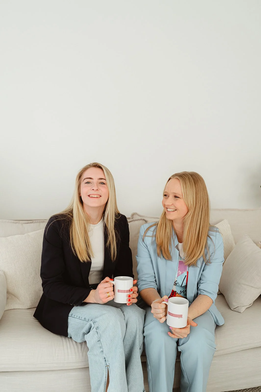 Twee vrouw zitten op een bank, lachen en drinken uit mokken.