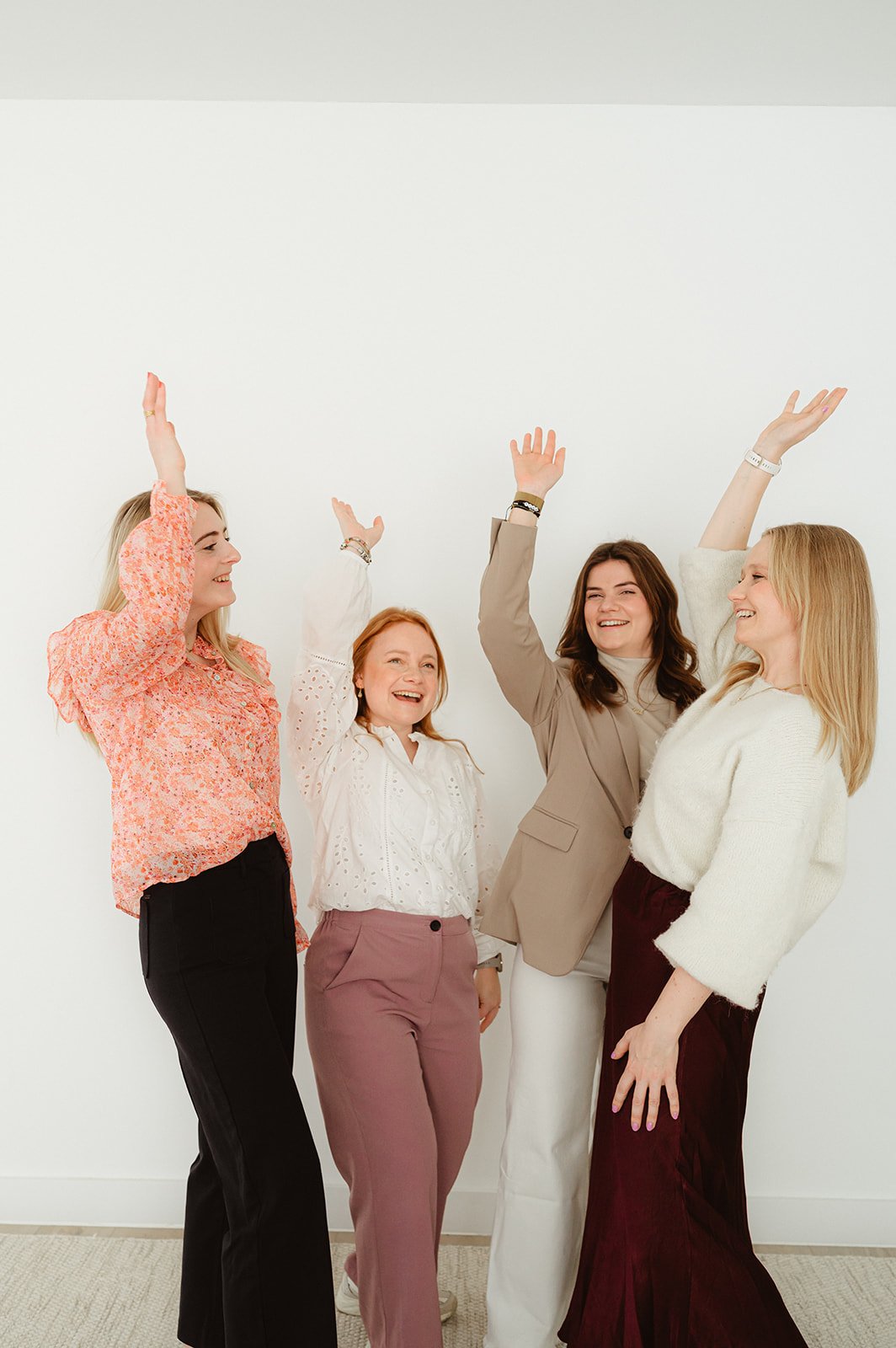 Vijf vrouwen die samen lachen en plezier maken terwijl ze met hun handen omhoog in de lucht wijzen, staand voor een witte muur.