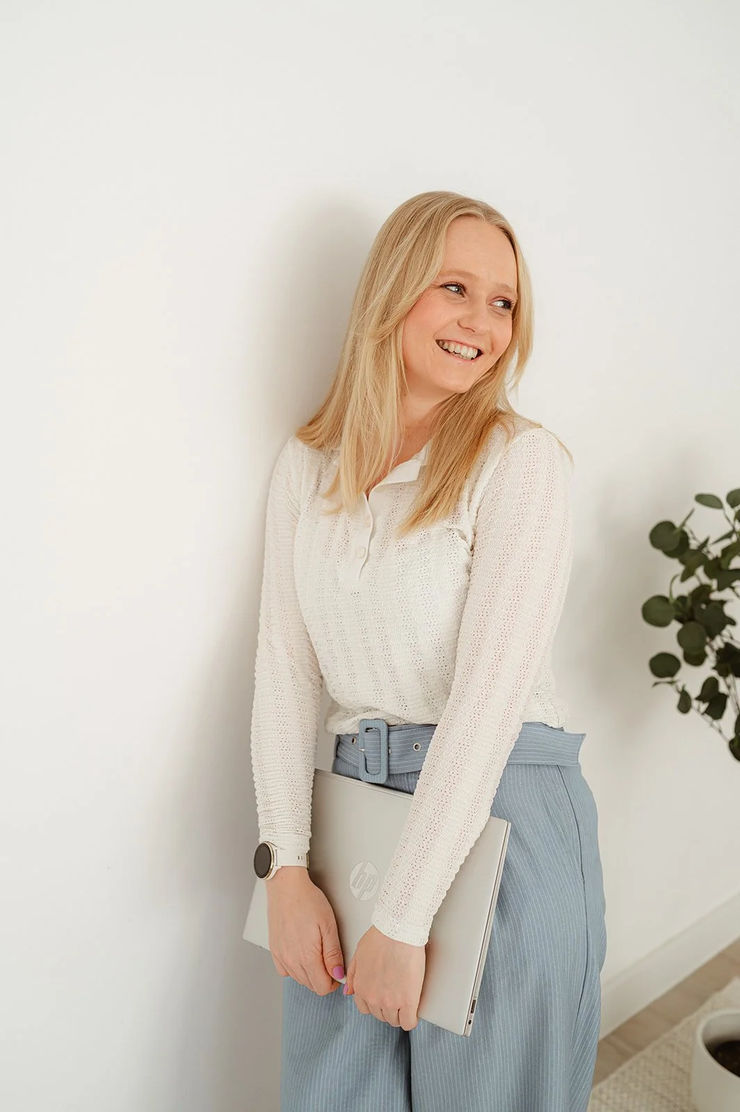 Een lachende vrouw met blond haar, gekleed in een witte trui en lichtblauwe broek, houdt een silverkleurige HP laptop en staat naast een witte muur met een plant op de achtergrond.