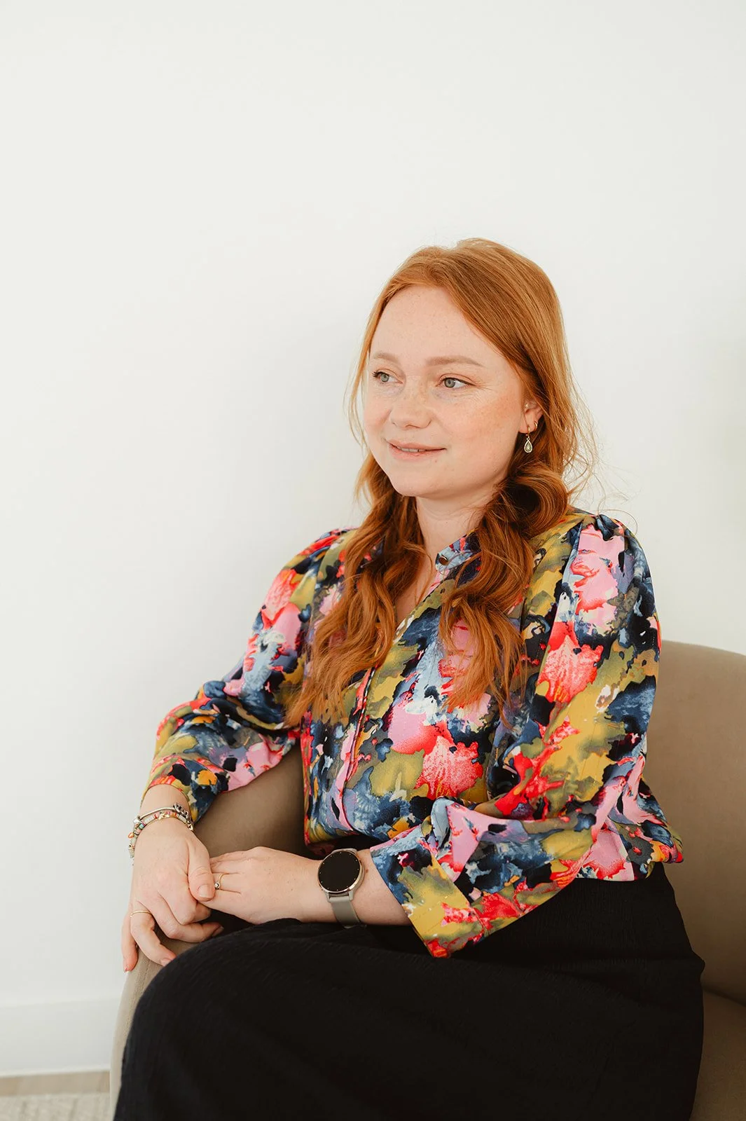 Vrouw met rood haar in een bloemenprint blouse, zittend op een bank, met een witte achtergrond.