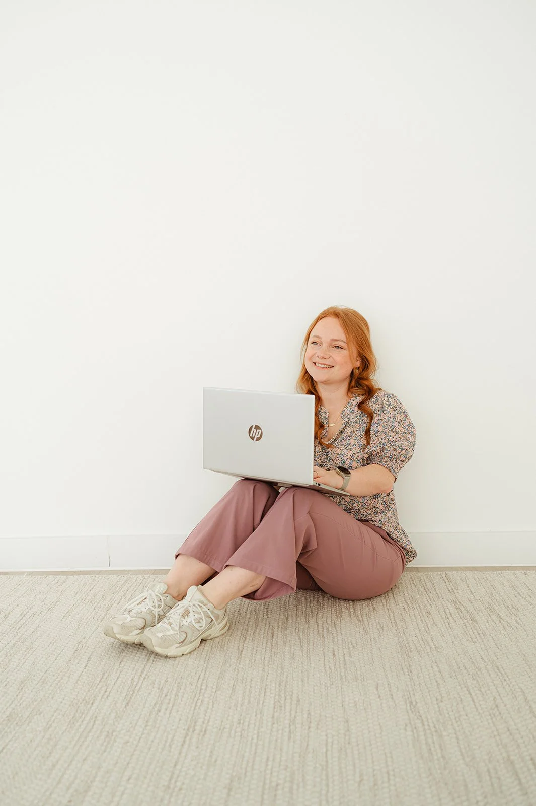 Een vrouw met rood haar zit op de grond tegen een witte muur, houdt een open HP laptop op haar schoot, draagt een bloemenblouse, roze broek, sneakers en kijkt tevreden.
