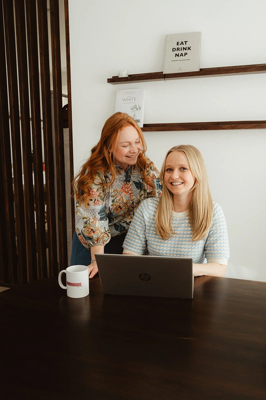 Twee vrouwen werken samen aan een laptop in een modern kantoor, lachen en lijken plezier te hebben.