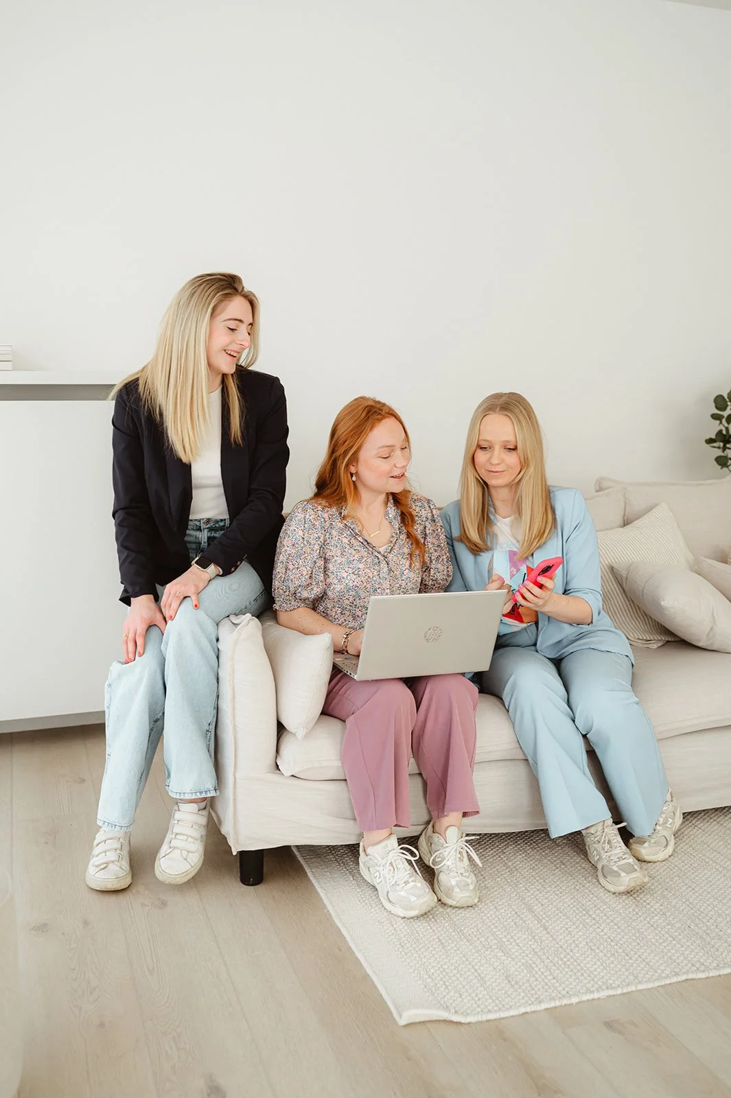 Dri vrouwen zitten op een bank in een woonkamer, kijken samen naar een laptop; twee meisjes gebruiken een smartphone.