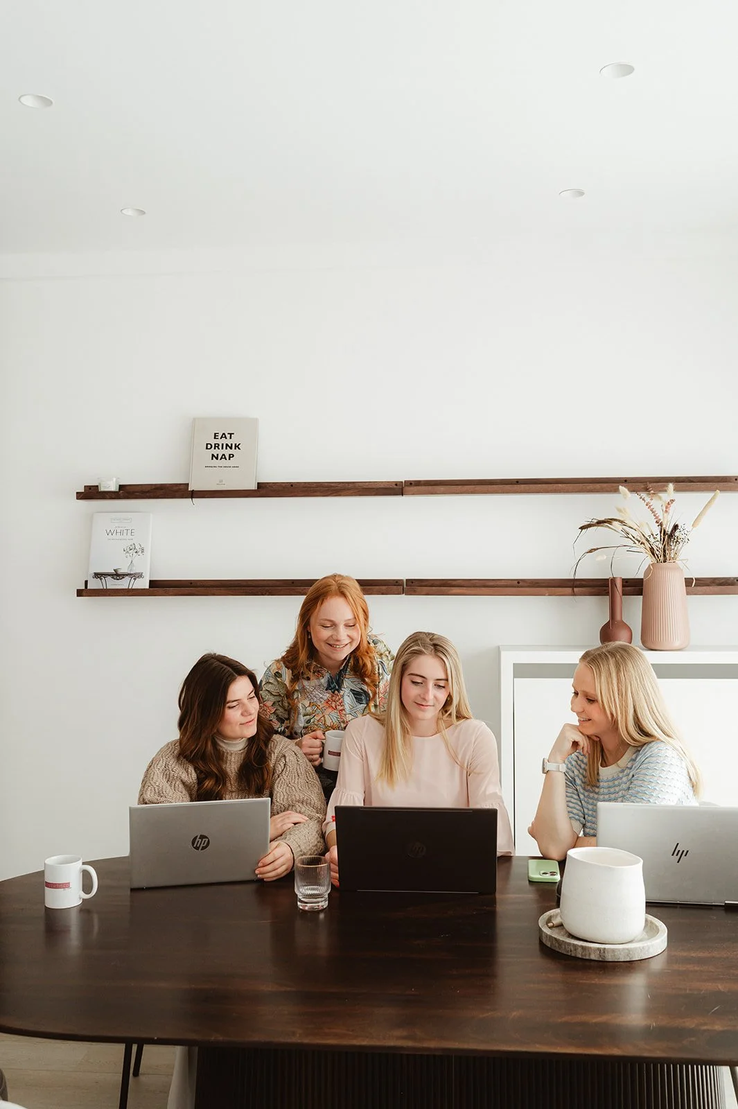Vijf vrouwen werken samen aan laptops aan een houten tafel in een lichte kamer.