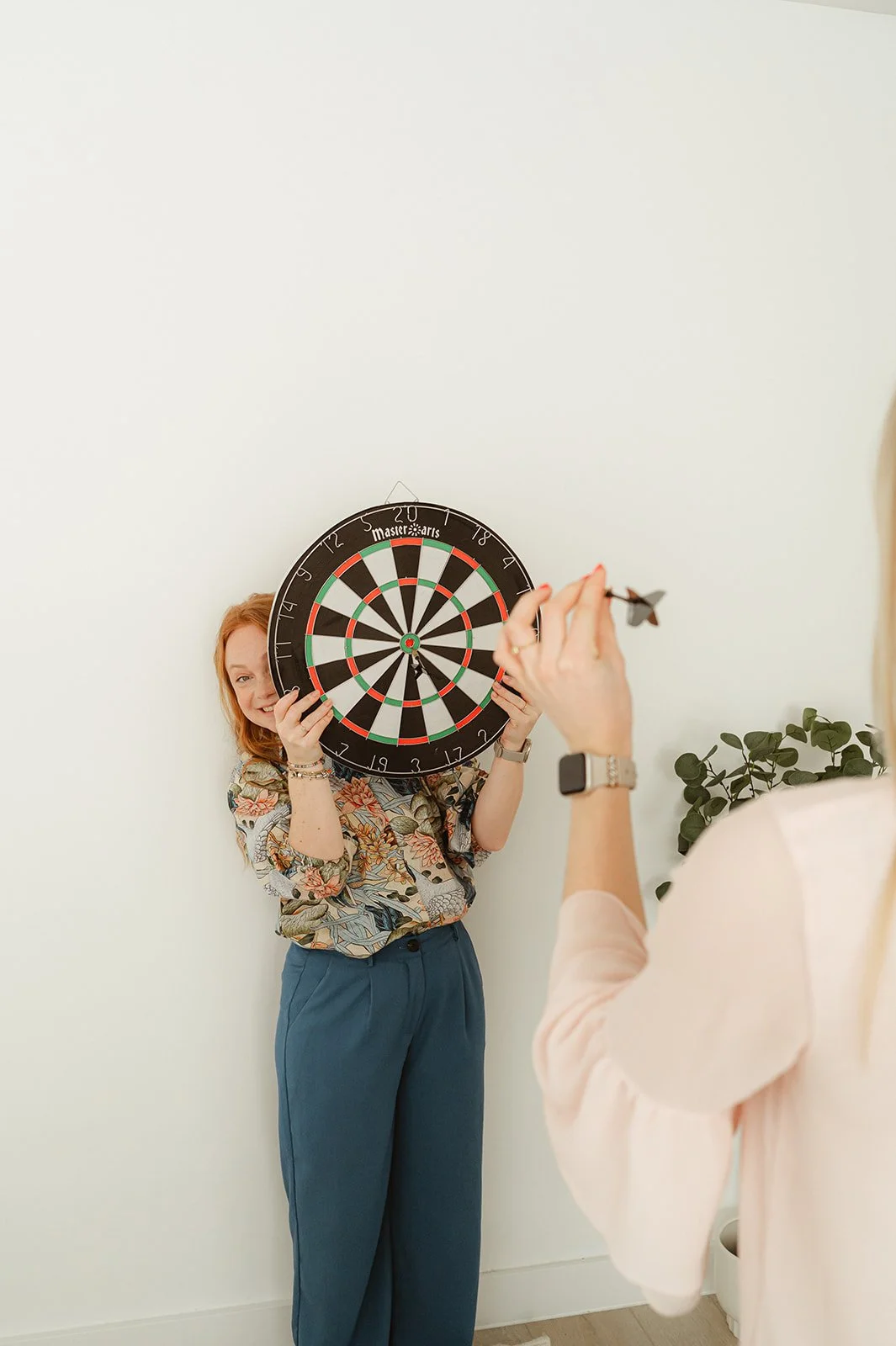 Vrouw houdt dartbord voor haar gezicht terwijl iemand anders klaarstaat om te gooien.