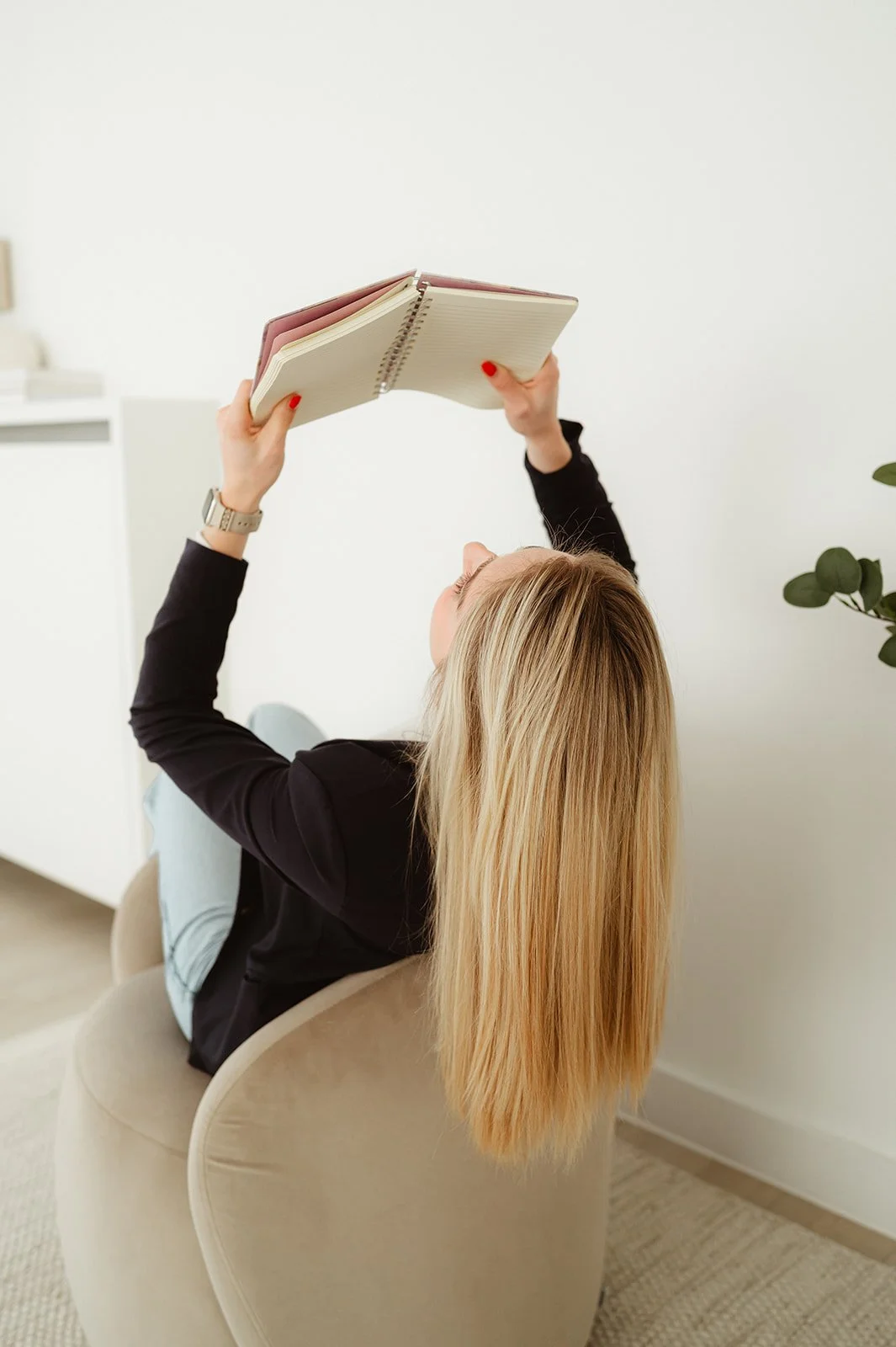 Vrouw ligt achterover in een beige stoel, leest een open notitieboekje en houdt het omhoog