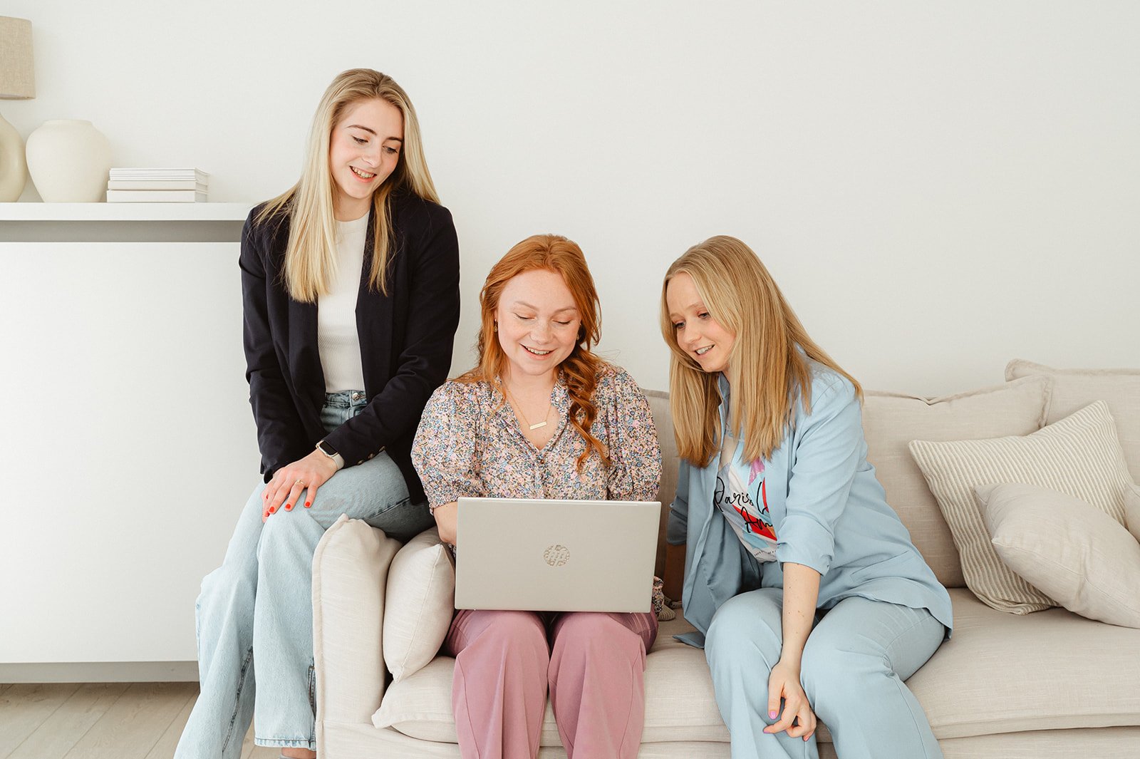Vrouw met rood haar zit op bank en gebruikt een laptop, omringd door twee vrouwen met blond haar die naar scherm kijken.