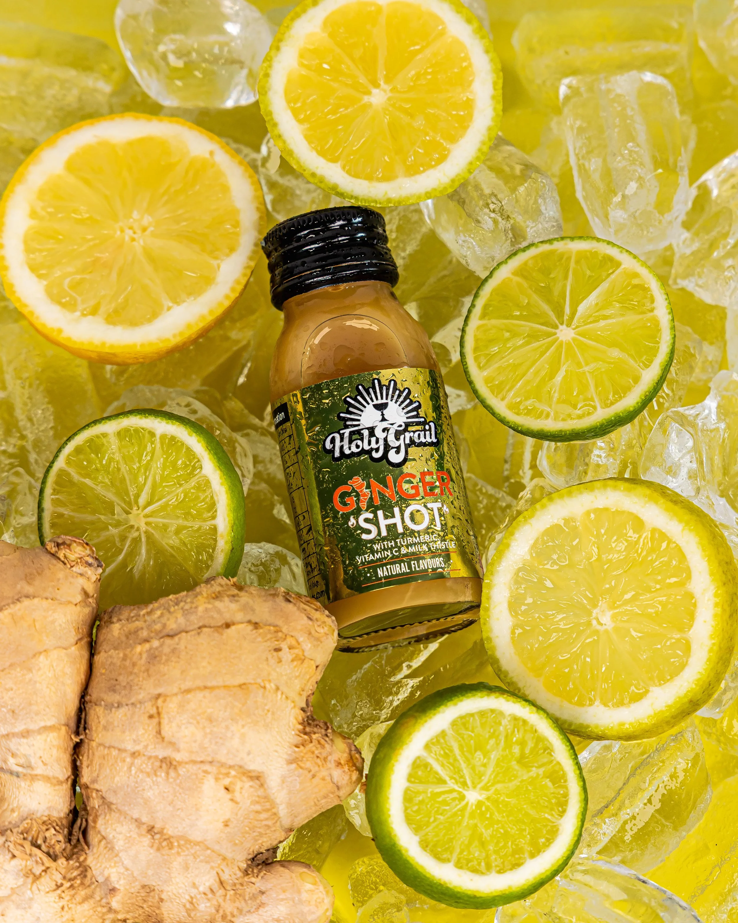 A bottle of Holy Grail Ginger Shot surrounded by lemon and lime slices, ginger root, and ice cubes.