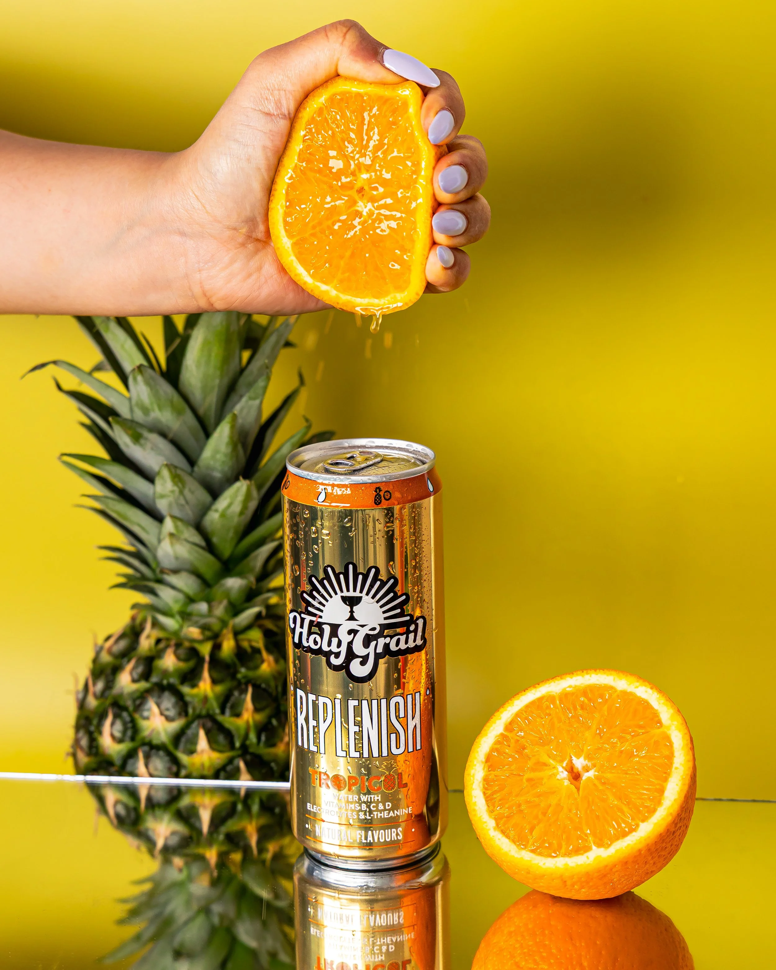 A hand squeezing juice from an orange half over a can of Holy Grail Replenishi Tropical drink, with a pineapple and a halved orange on a reflective surface against a yellow background.