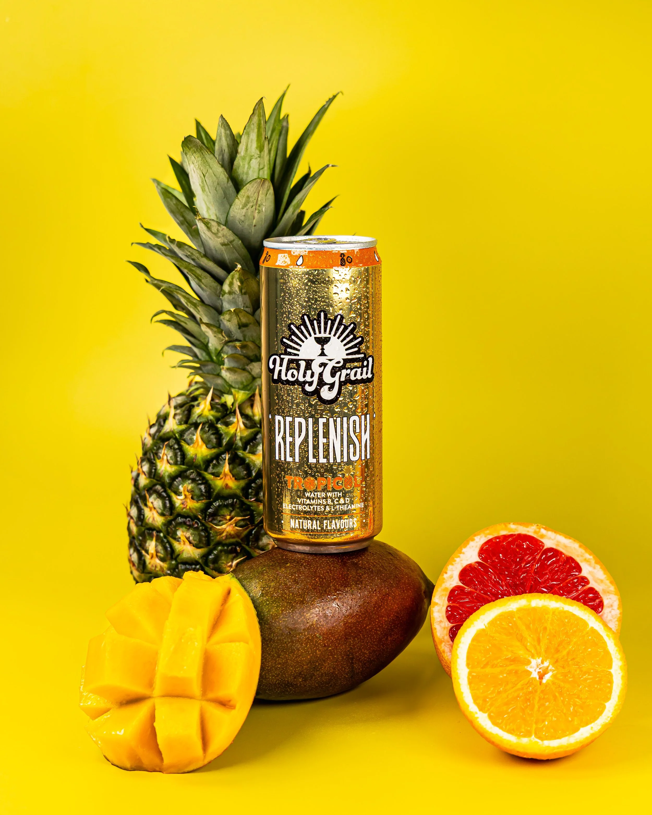 A pineapple, a mango, a grapefruit, and an orange along with a can of Holy Grail replenish tropical flavored drink on a yellow background.