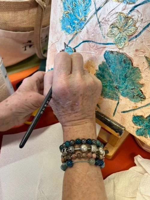 Une personne âgée peint une toile avec des fleurs colorées, en utilisant un pinceau, sur une surface en bois.