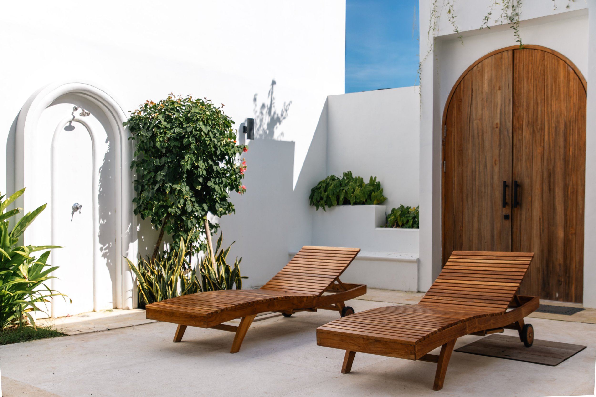 Outdoor patio with two wooden lounge chairs, a white wall with a wooden arched door, green plants, and a blue sky.