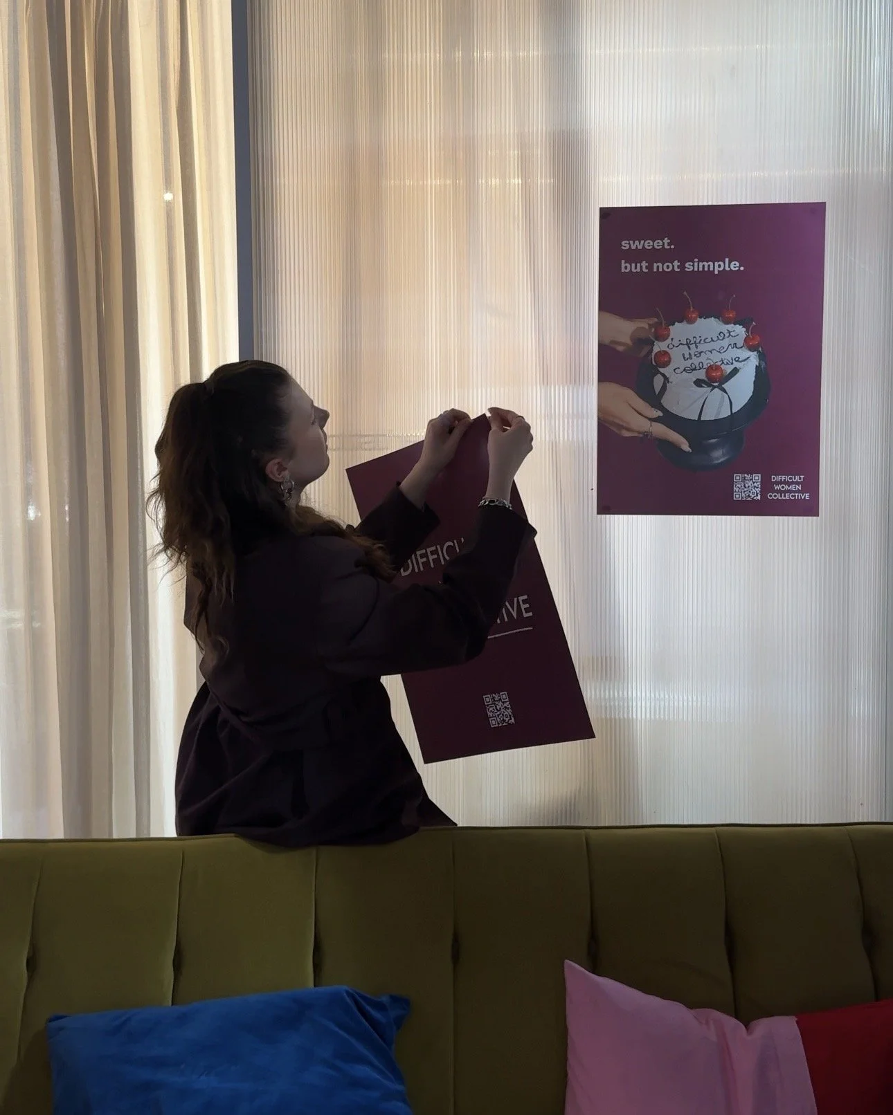 A woman attaching a maroon sign to a wall, with a poster nearby showing a teapot with cherries and the words 'sweet, but not simple.'
