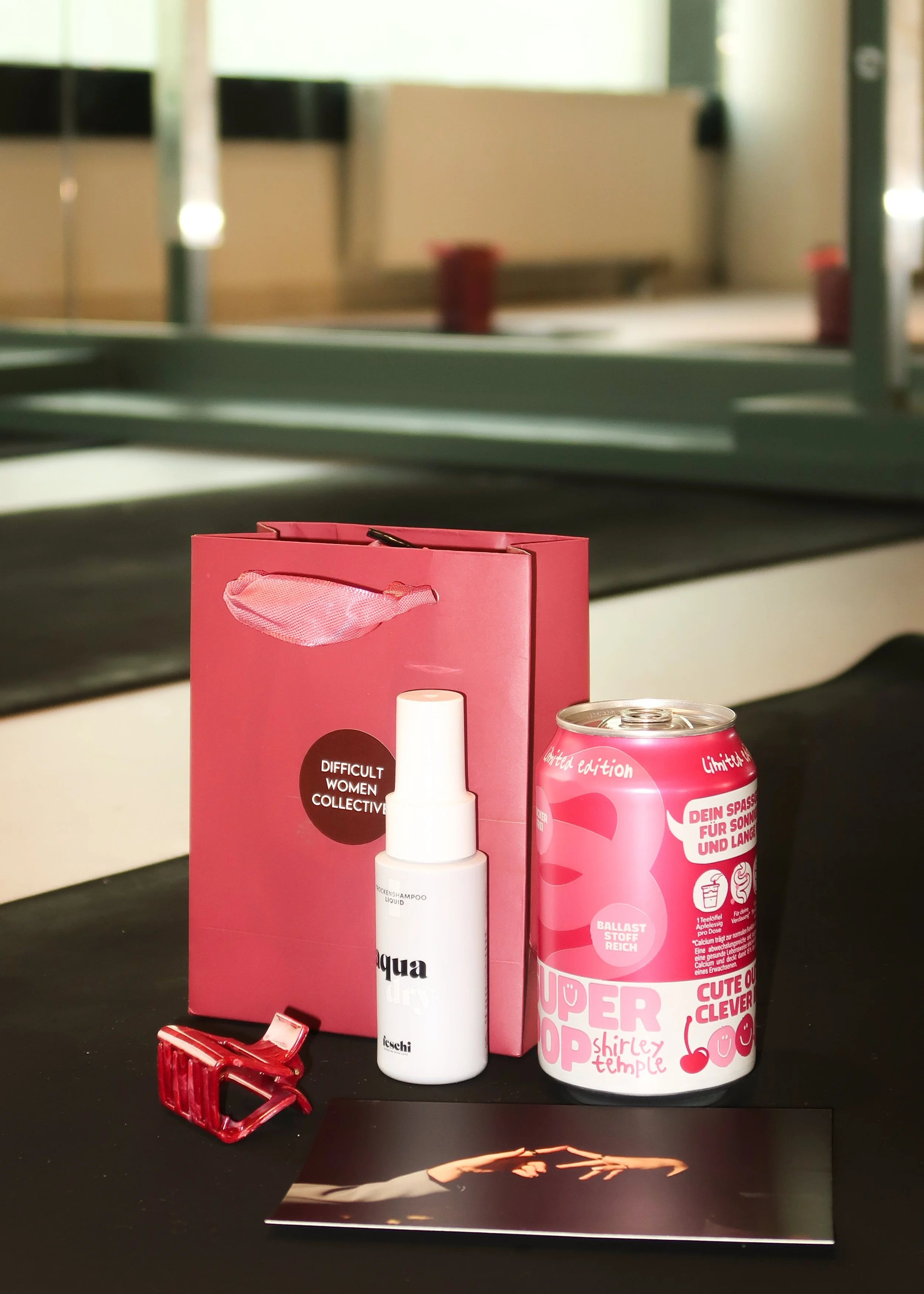 Close-up of various small personal care items on a black surface, including a pink gift bag, a white bottle, a red miniature purse, and a pink can with text, with a blurred background of a window and furniture.