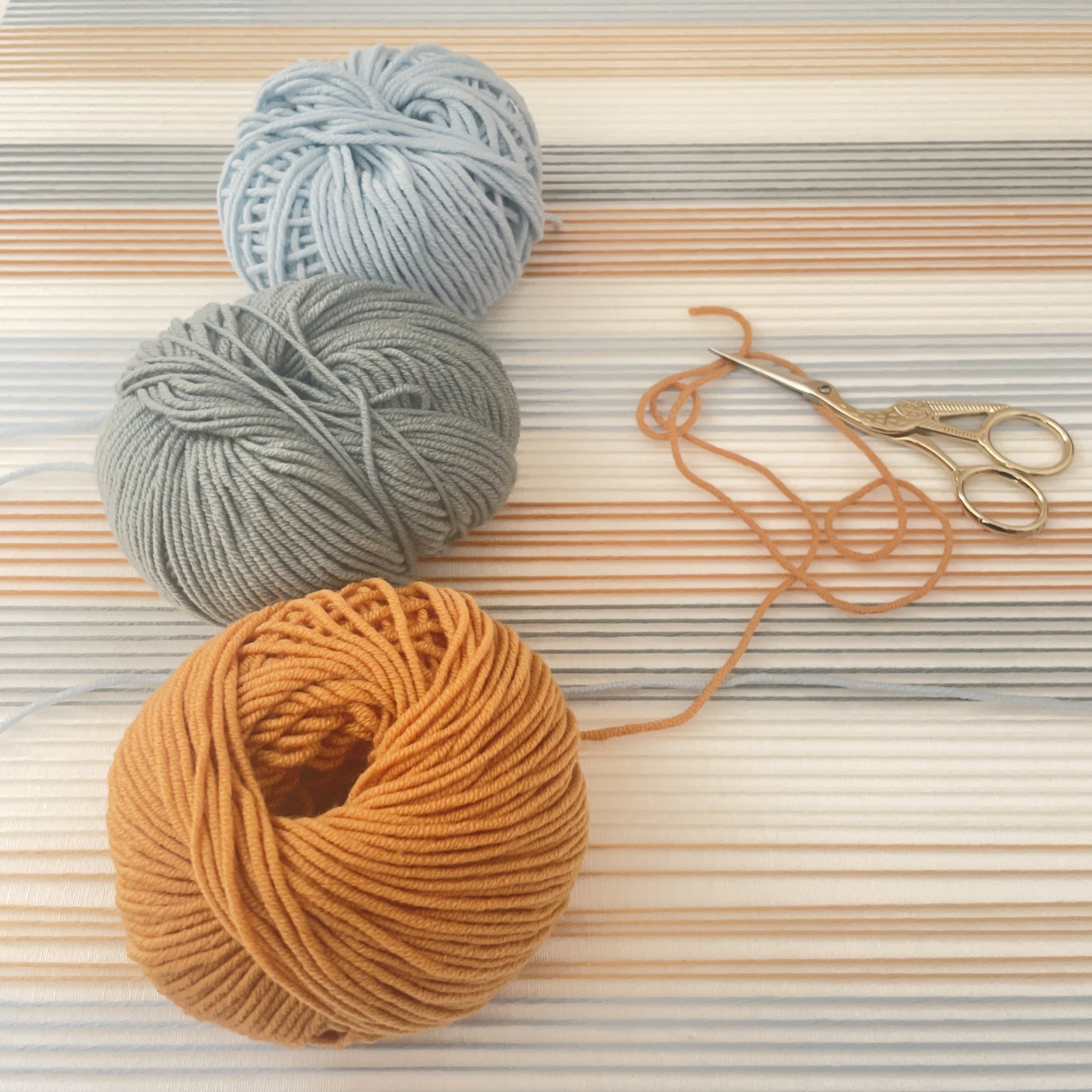 Pelotes de hilo en color gris, azul y naranja, junto con tijeras de coser doradas, sobre una superficie de piso de madera con rayas blancas y beige.
