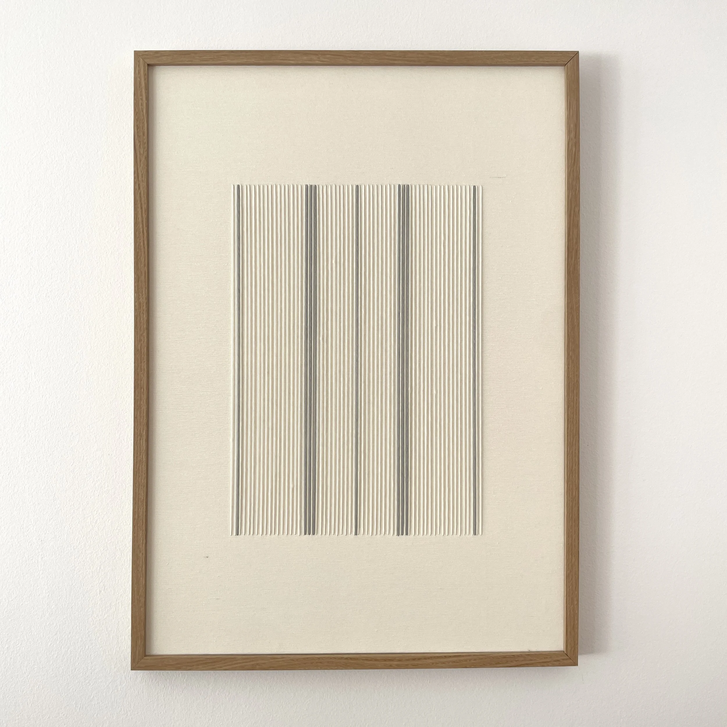 Cuadro con marco de madera y una parte de una obra con líneas verticales en relieve en tonos grises y blancos, colgado en una pared blanca.
