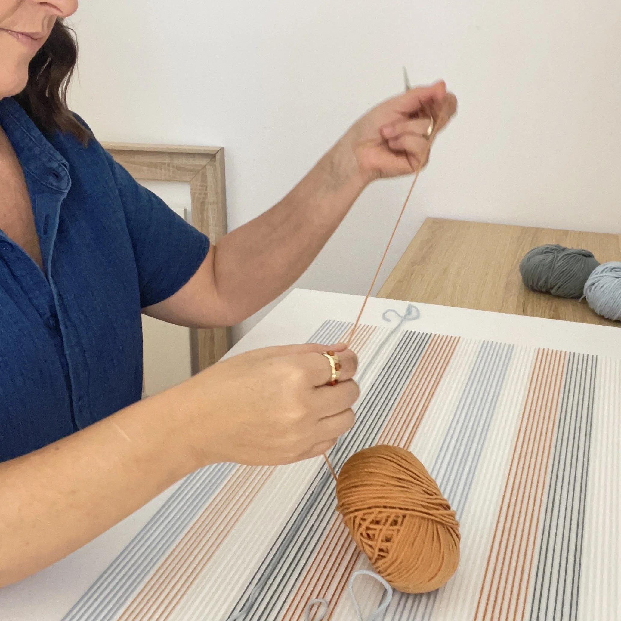 Una mujer con blusa azul teje con hilo de color naranja y gris sobre una mesa de madera, mientras hay bobinas de hilo a su lado.