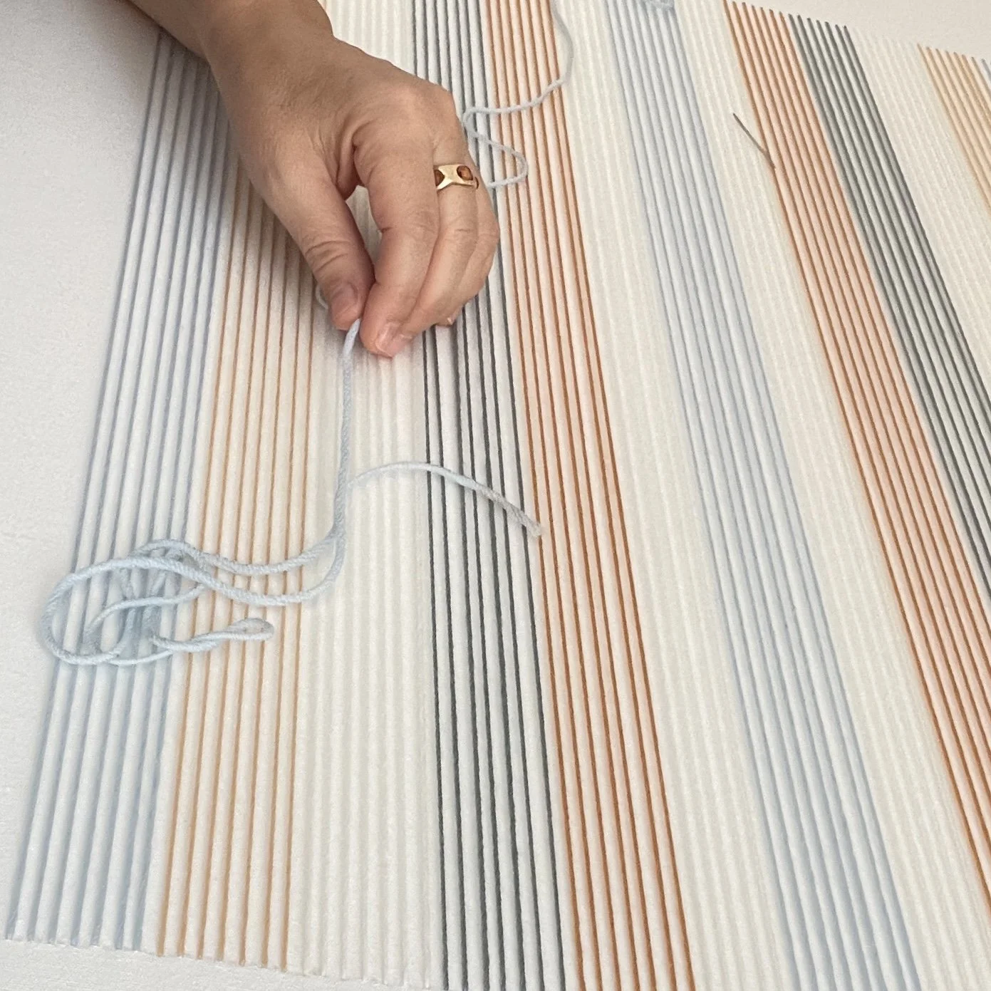 Una mano colocando hilos en un tapete con líneas de colores beige, gris y naranja.