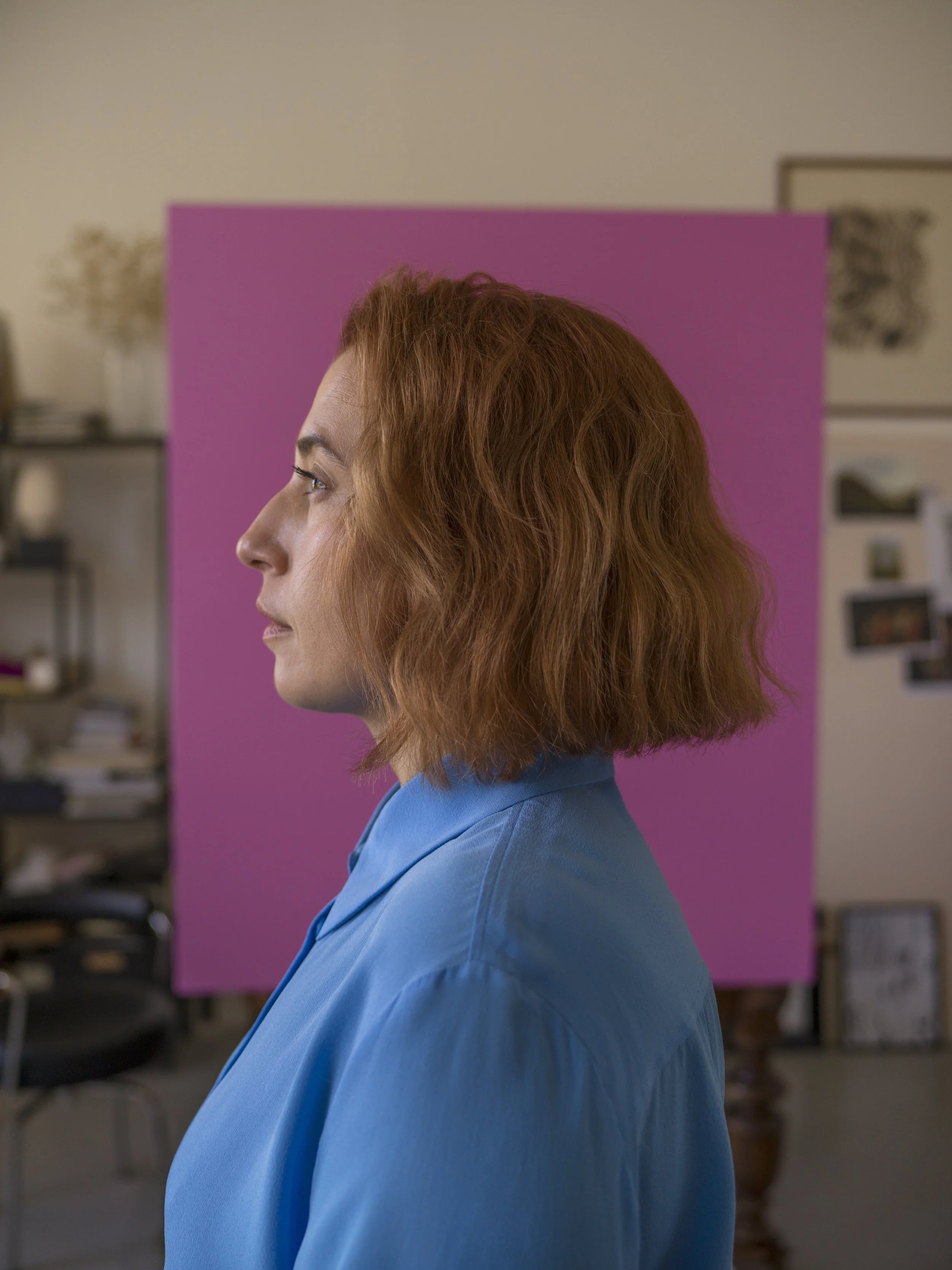 Profile of a woman with red hair wearing a blue shirt, standing indoors with a pink backdrop behind her.