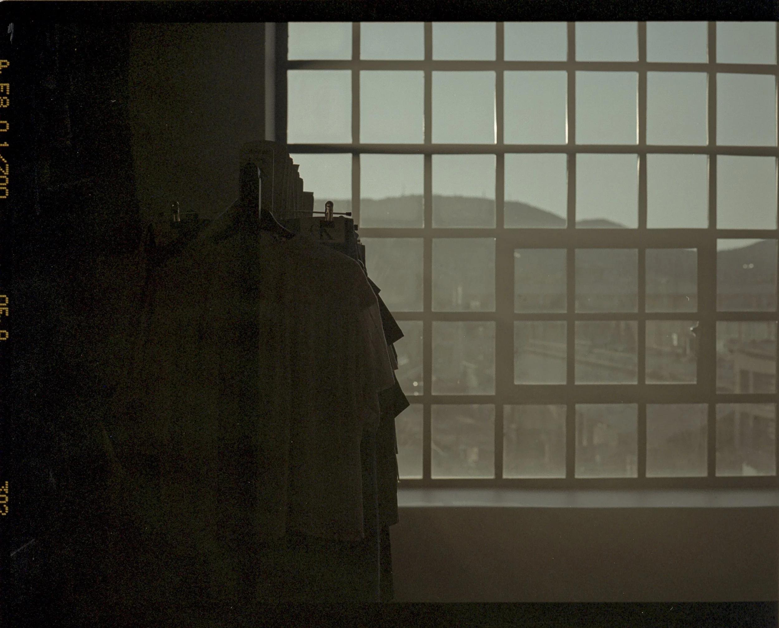 Clothes hanging on a rack in front of a large grid window with a cityscape and mountains in the background, viewed during daylight hours.