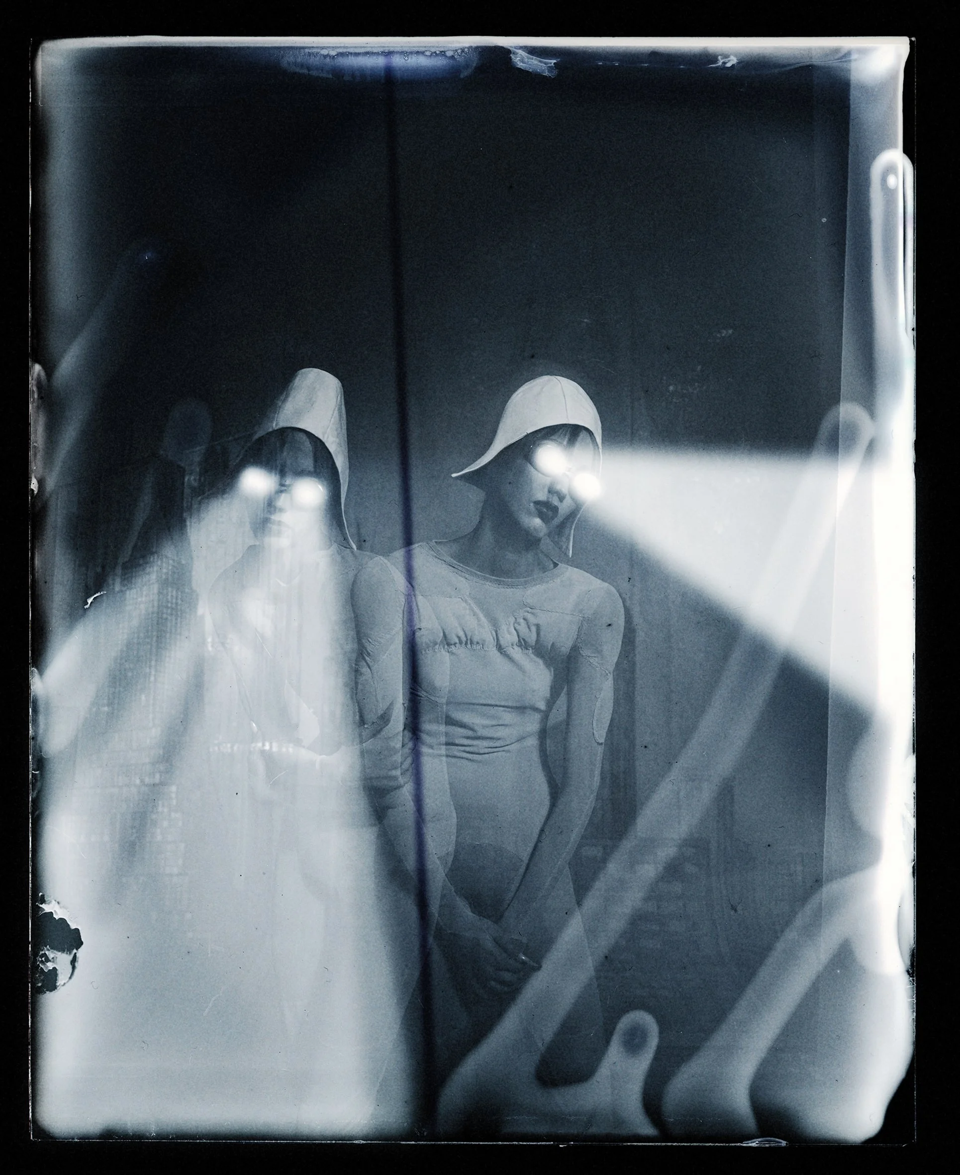 Black and white photo of three women with glowing eyes, wearing hoods and vintage clothing, standing in a dark room with light reflecting off a glass or plastic surface in front of them.