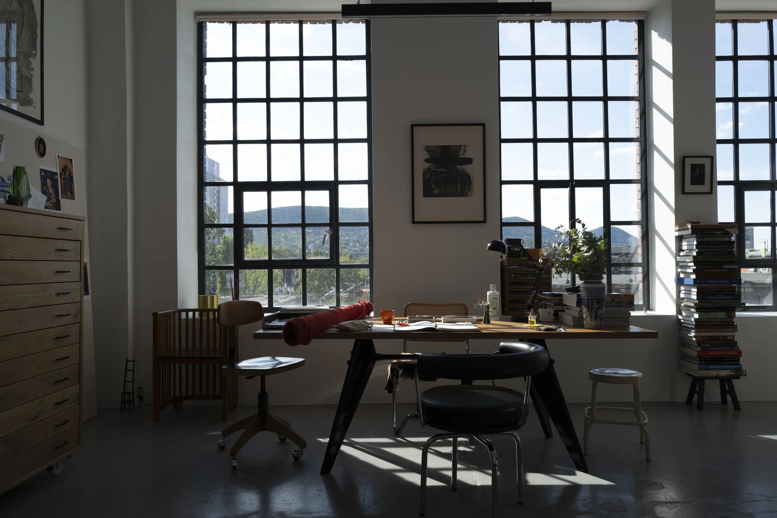 Bright office with large grid windows, a wooden desk with various items including books, plants, and a lamp, surrounded by chairs and storage units.